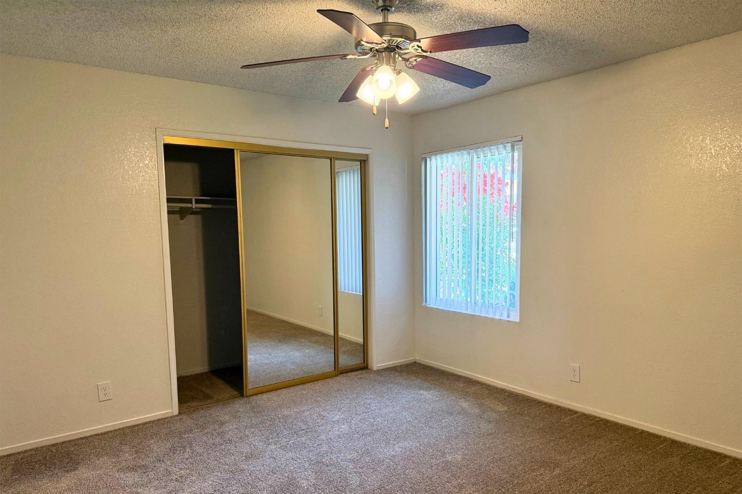 A well-lit room featuring light-colored walls and carpeted flooring. It has a ceiling fan with three lights, a large mirrored closet with sliding doors, and a window with vertical blinds allowing natural light to enter. The space is empty, creating a clean and inviting atmosphere.