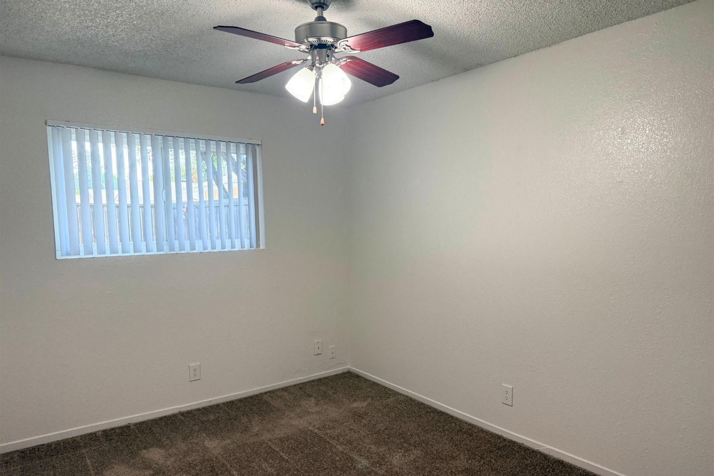 Empty room featuring a ceiling fan with four blades, a window with vertical blinds allowing natural light, and light-colored walls. The floor is carpeted. The space appears clean and freshly painted, suitable for various uses.