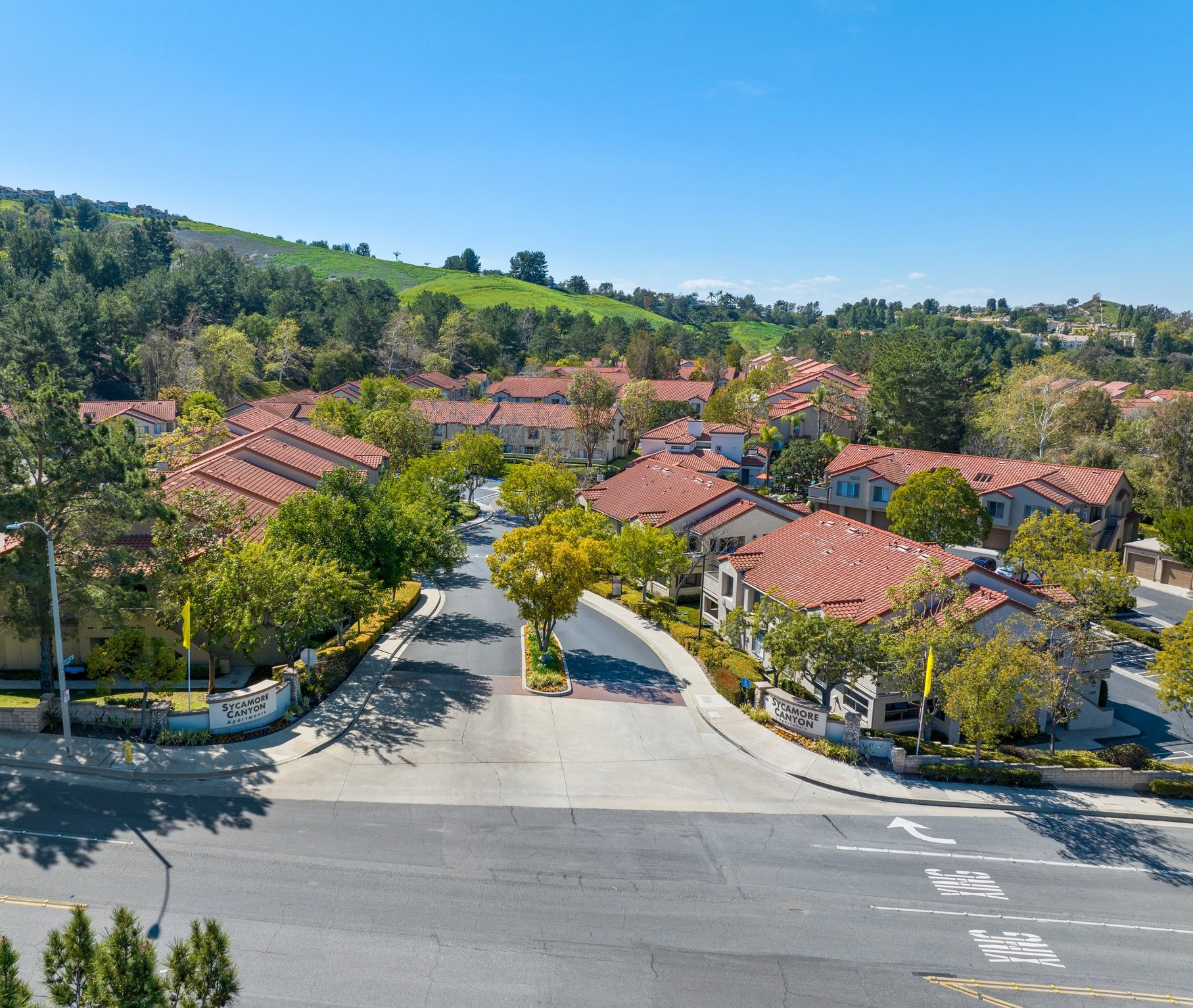 Aerial view of a suburban neighborhood featuring several residential buildings with reddish-tiled roofs, surrounded by trees and greenery. The streets are well-maintained, and the landscape includes hills in the background under a clear blue sky.