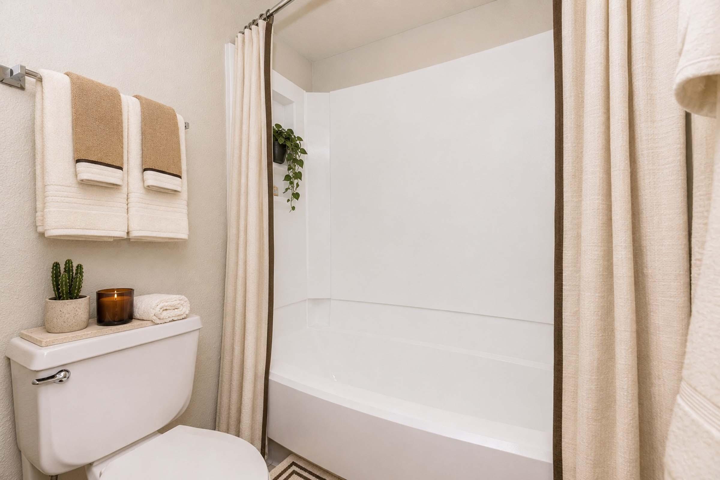 A modern bathroom featuring a bathtub with a shower curtain, neatly arranged towels on a rack, a small potted plant, a scented candle, and a decorative towel on the toilet tank. The walls are painted a light color, creating a clean and inviting atmosphere.