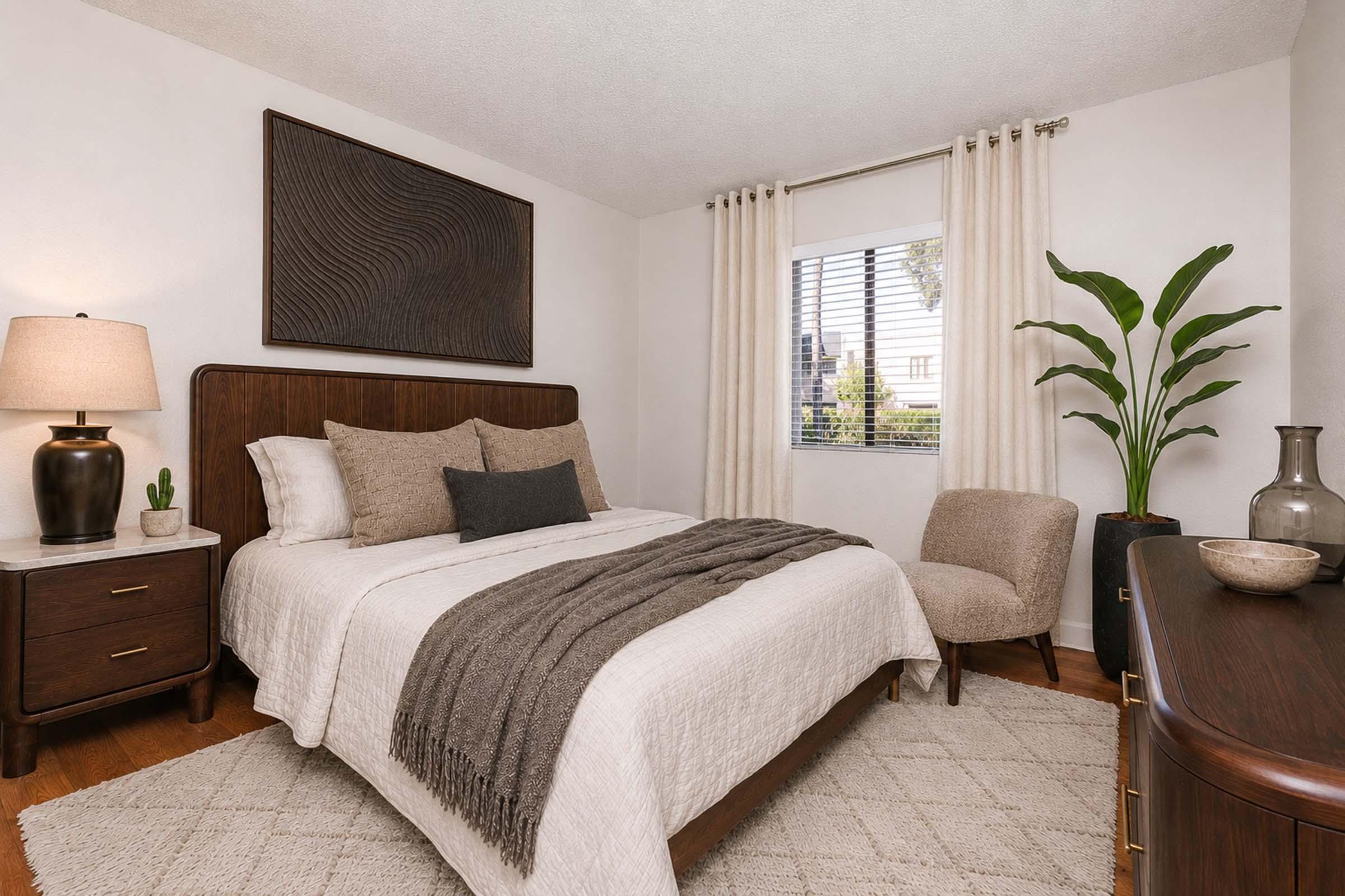 A cozy bedroom featuring a wooden bed with a light-colored duvet and decorative pillows. There's a stylish lamp on a nightstand, a plant in the corner, and a window letting in natural light. A vase and chair add to the modern decor, complemented by a textured wall art piece above the bed.