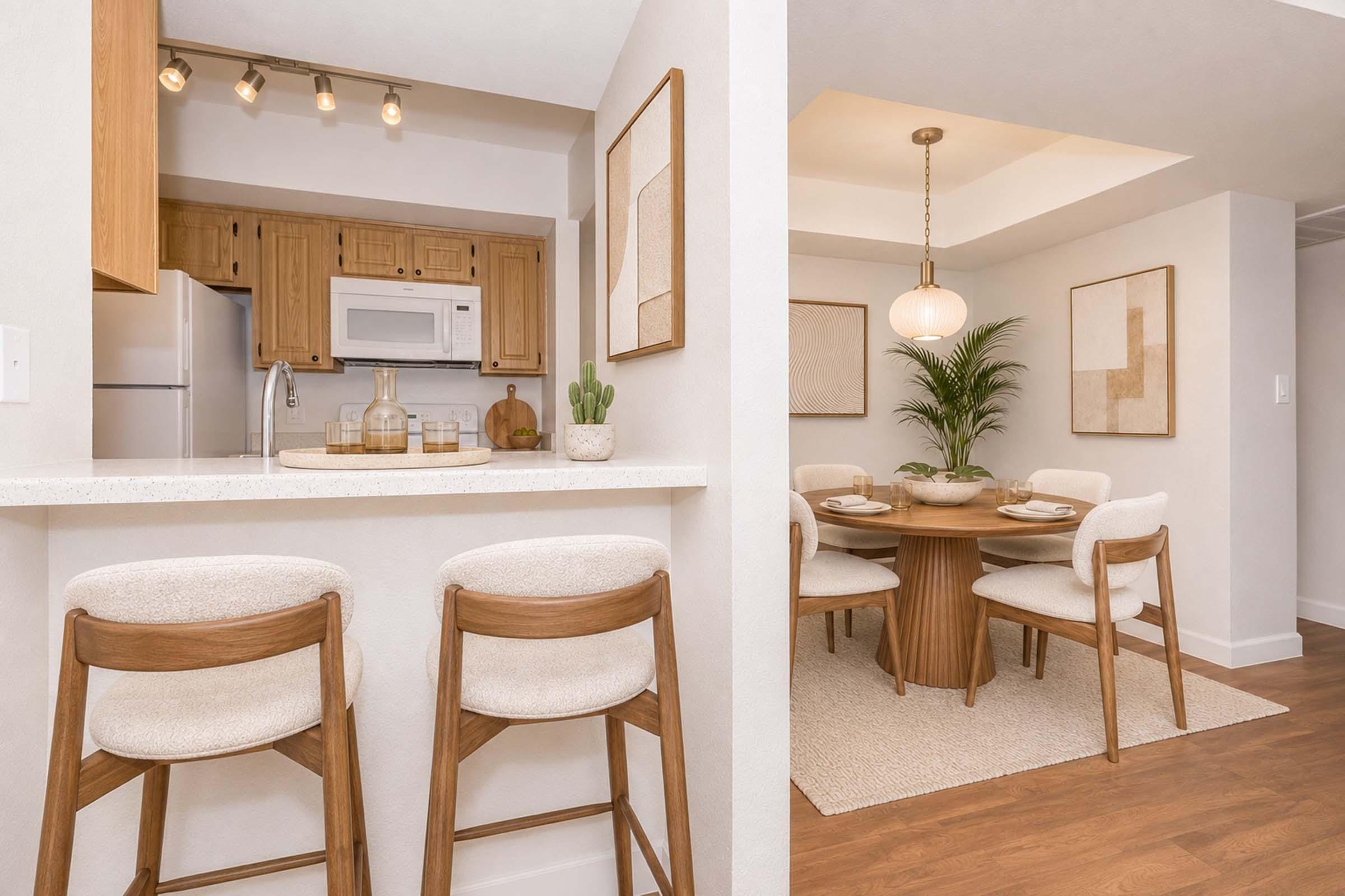 Modern kitchen and dining area featuring a light wooden bar with two stools, an island with decorative items, and a round dining table set for four. The space is decorated in neutral tones with framed artwork on the walls and a plant in the corner, creating a warm and inviting atmosphere.