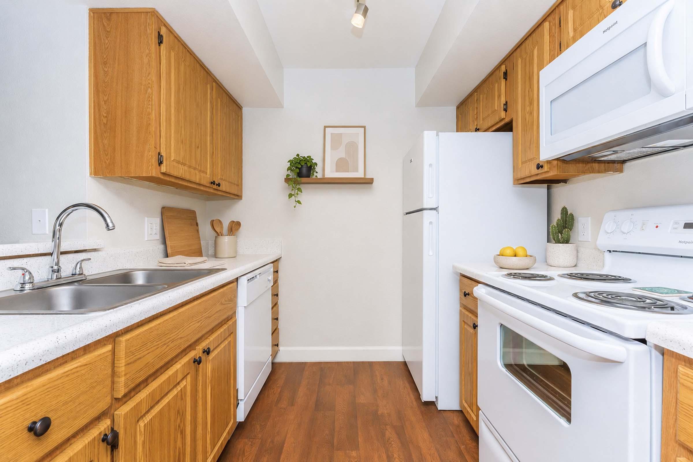 A modern kitchen featuring wooden cabinets, white countertops, and stainless-steel appliances. The layout includes a double sink, a dishwasher, and a refrigerator. There is a shelf with a small plant and framed artwork on the wall, along with a bowl of oranges on the counter, creating a warm and inviting atmosphere.
