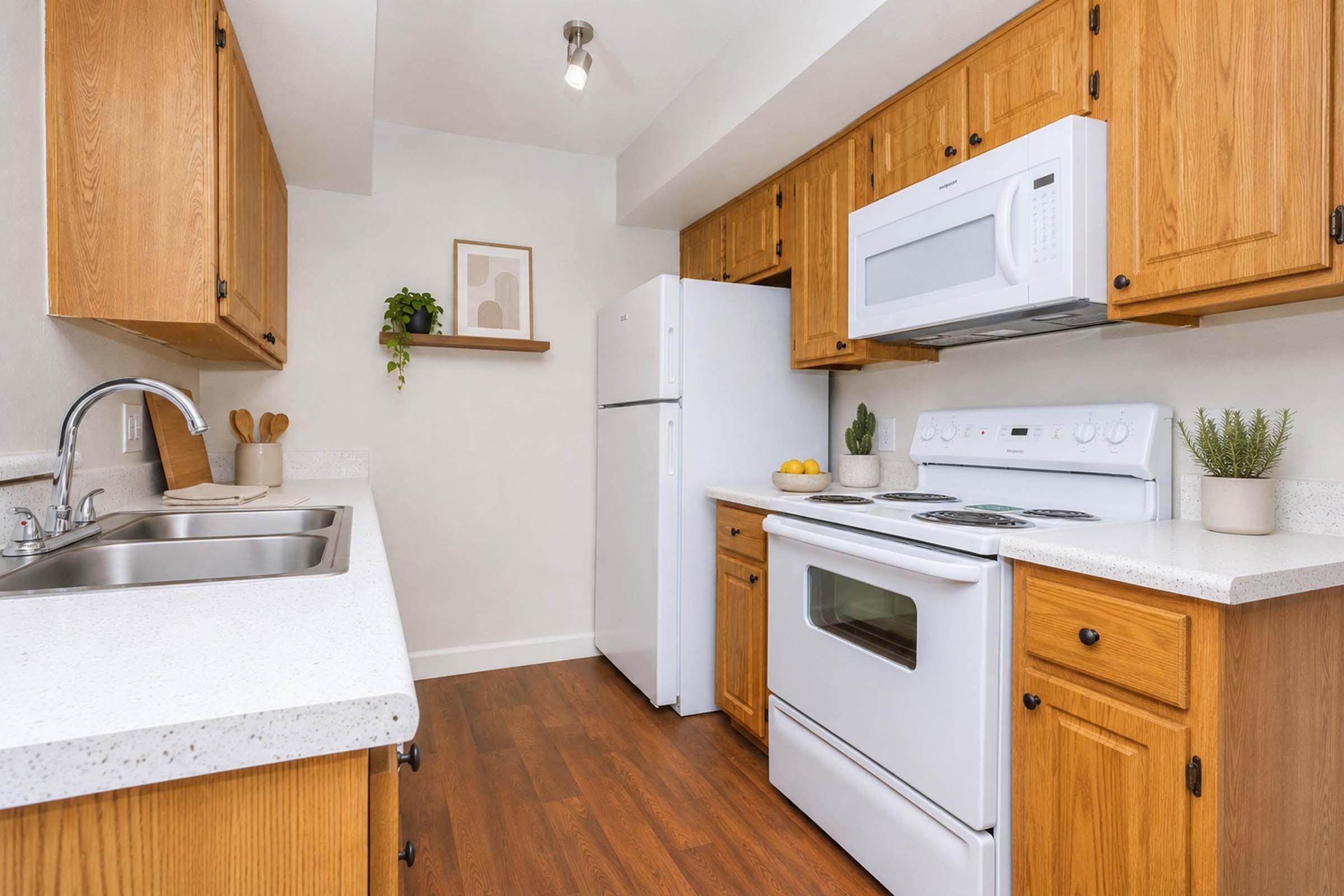 A modern kitchen featuring wooden cabinets, a white refrigerator, and stove. There is a stainless steel sink on the left, a countertop with a few kitchen items, and some greenery. The walls are light-colored, creating a bright and airy atmosphere.