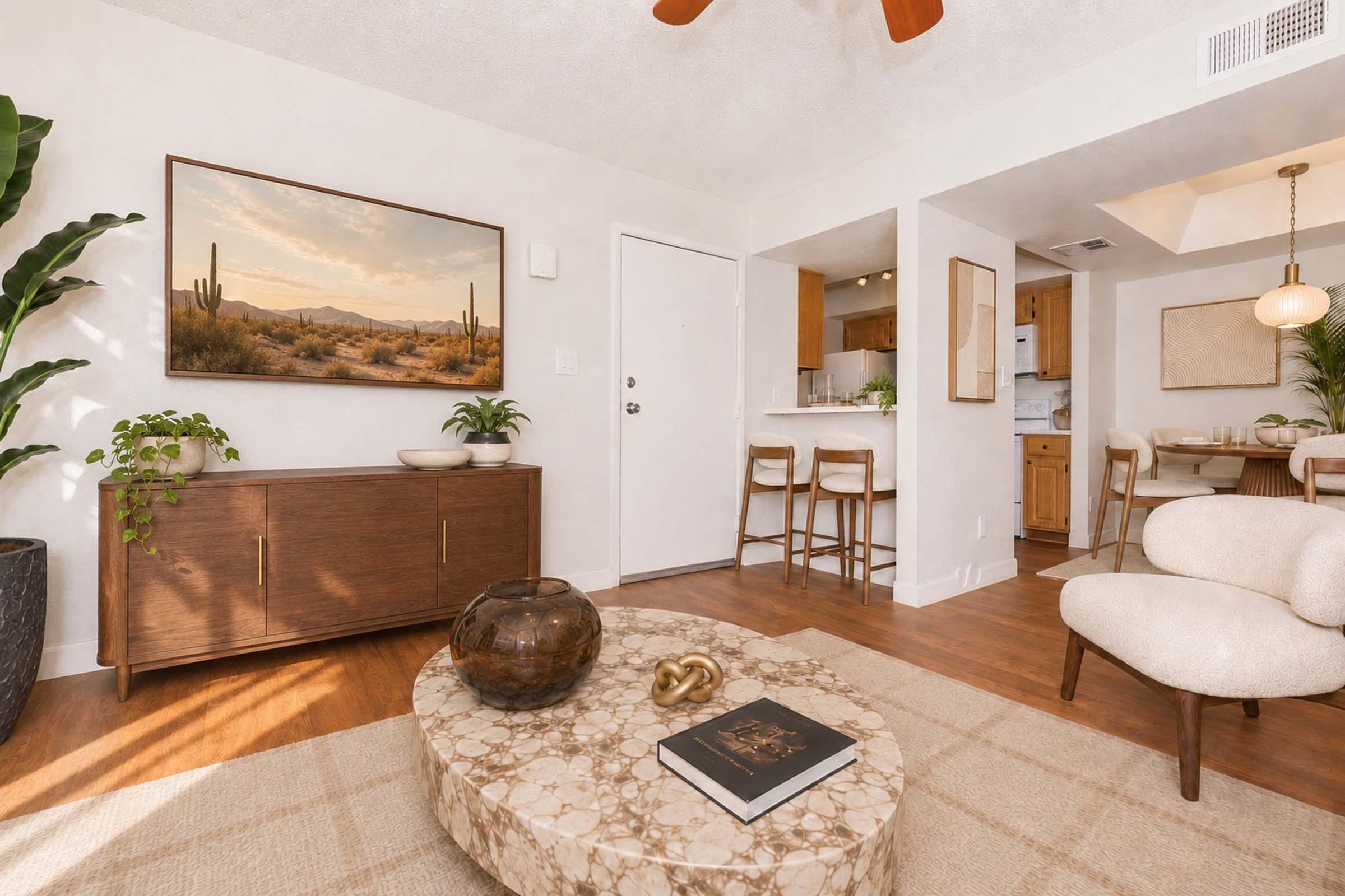 A modern living room featuring a cozy seating area with a round coffee table, a minimalist cabinet, and a framed landscape painting of cacti. The space includes plants for decoration and leads to an open kitchen area with bar stools. Soft lighting enhances the warm, inviting atmosphere.