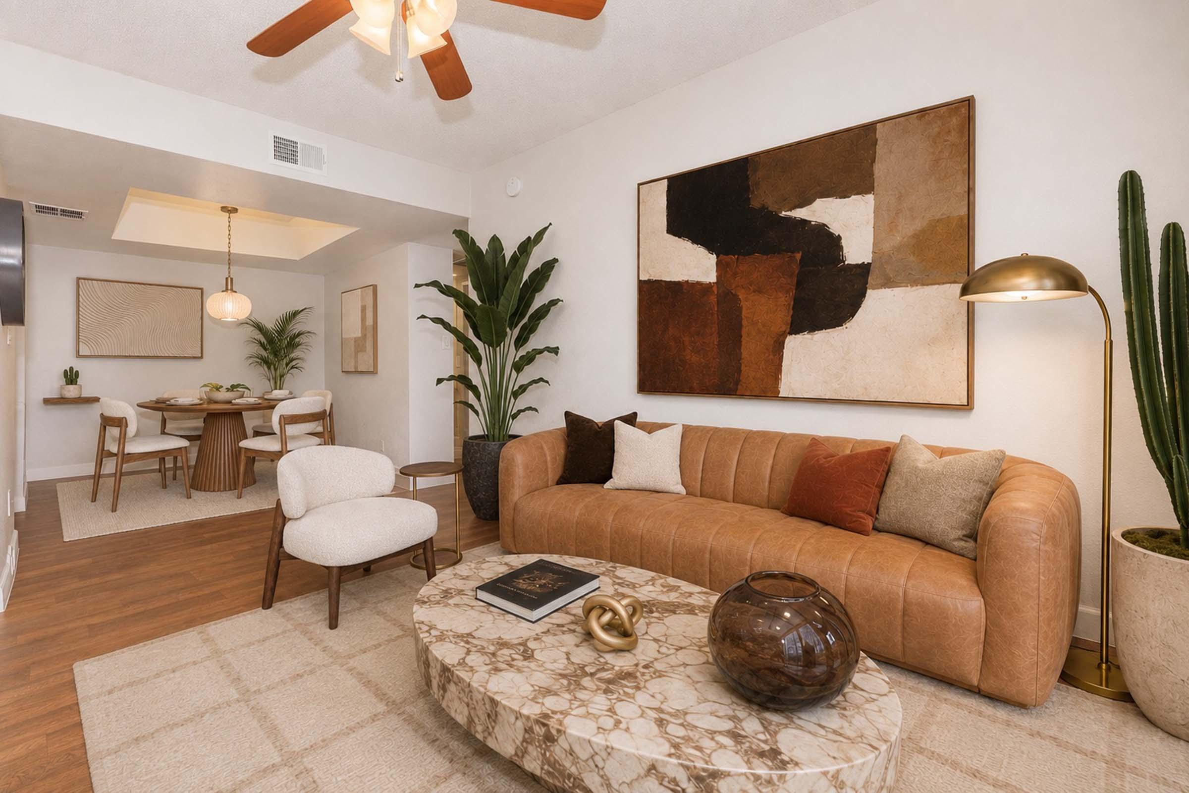 A stylish living room featuring a brown leather sofa with decorative pillows, a round marble coffee table, and a large abstract wall art piece. In the background, there's a dining area with a wooden table and chairs, along with potted plants adding greenery to the space. The room has a warm and inviting atmosphere.