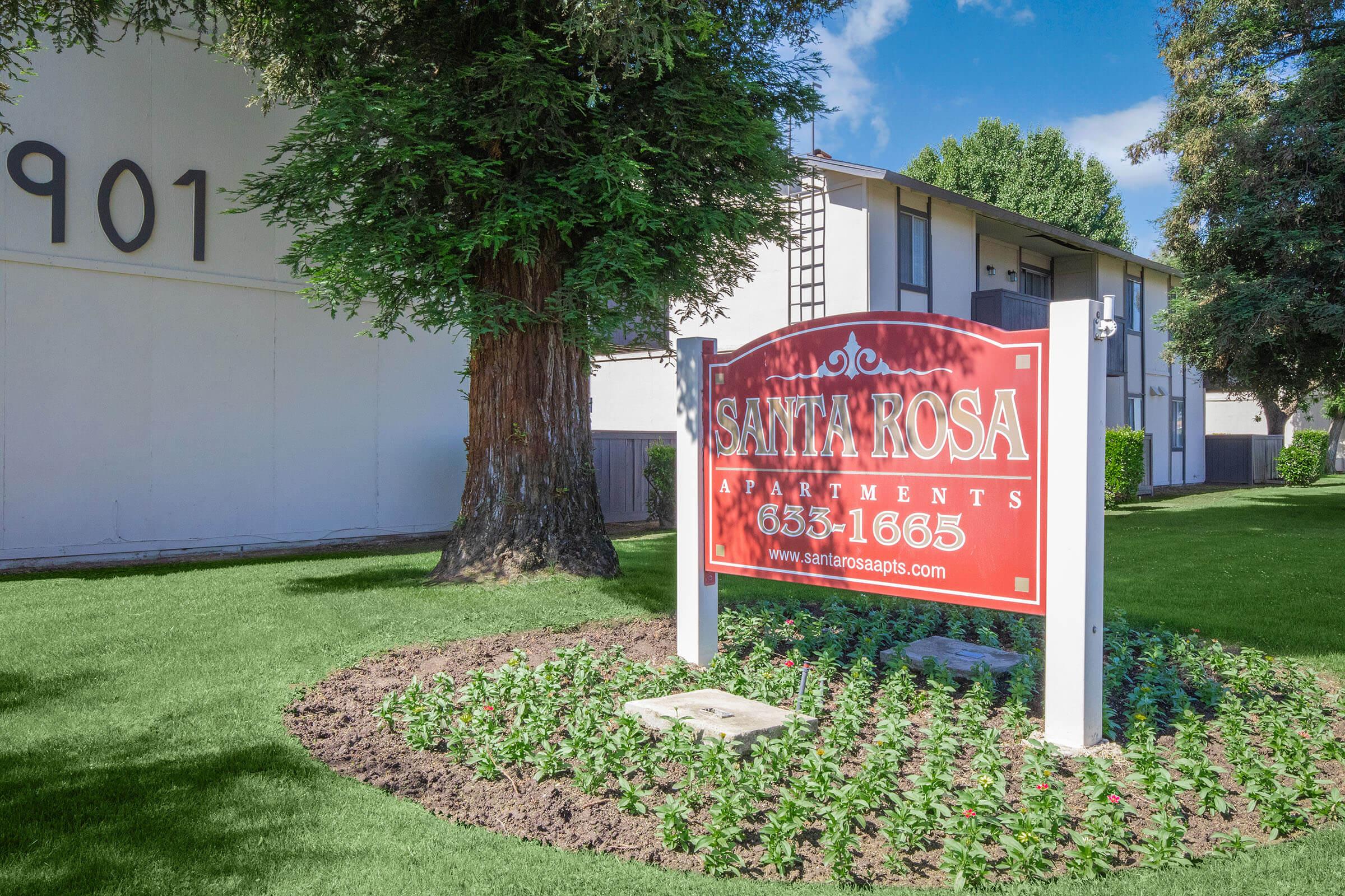 Sign for Santa Rosa Apartments with contact number and website, surrounded by well-maintained flower beds and a large tree, set against a clear blue sky. The apartment complex appears clean and inviting.