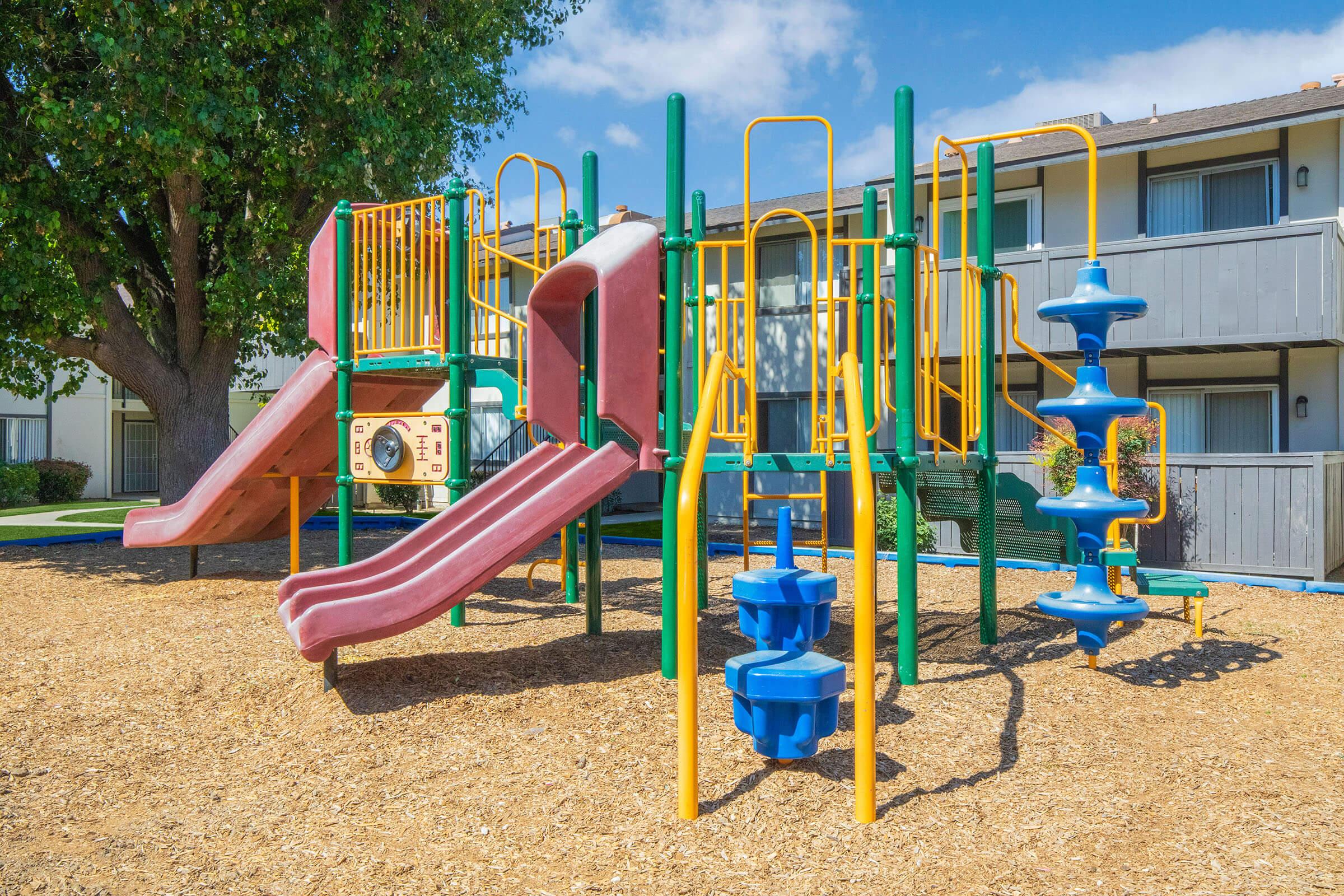 Colorful playground equipment featuring two slides, climbing structures, and a spiral slide, surrounded by mulch. The setting includes nearby apartments and green trees under a clear blue sky, providing a vibrant and inviting atmosphere for children to play.
