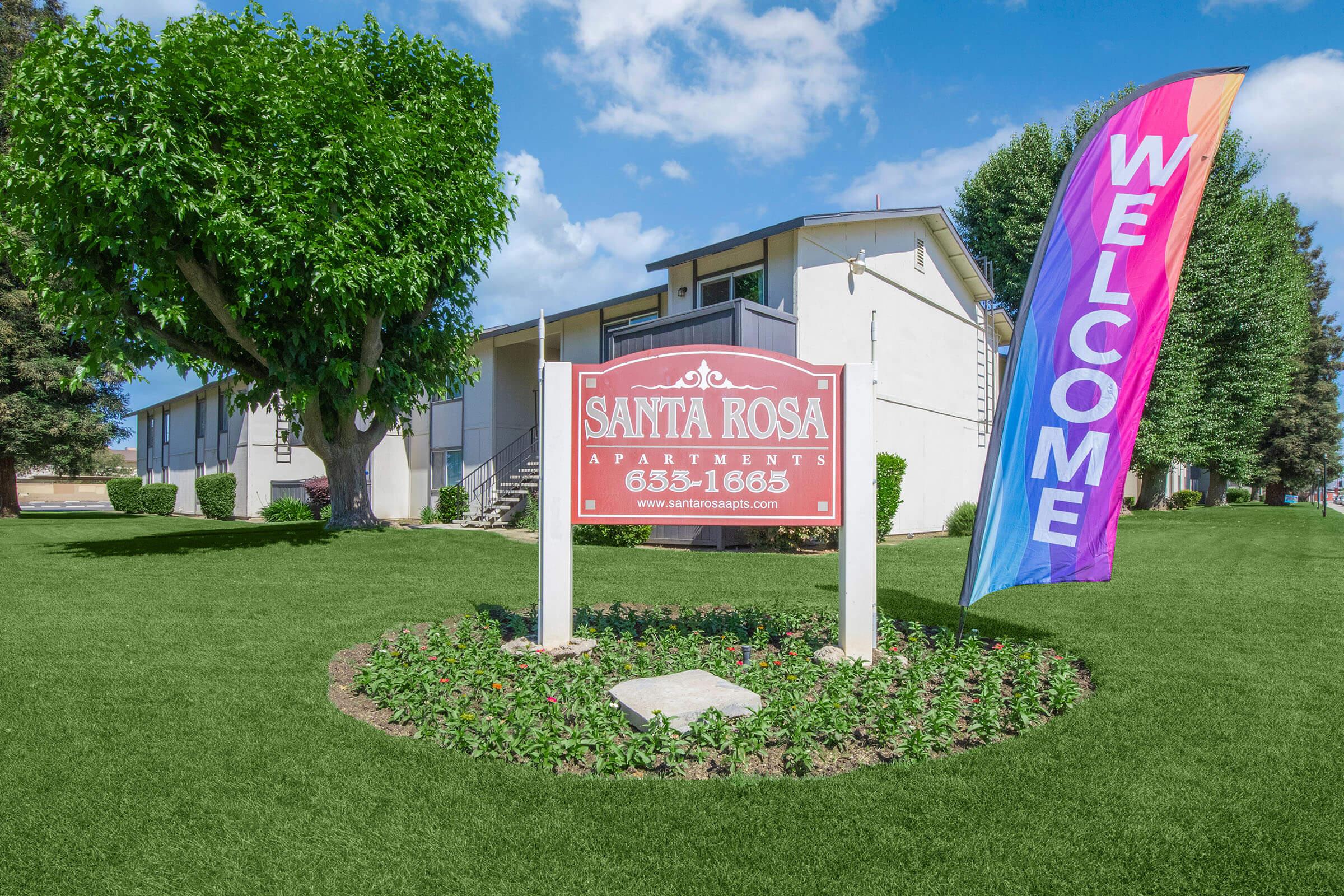 A well-maintained apartment complex sign reading "Santa Rosa Apartments" with a contact number. In front of the sign, a colorful "WELCOME" banner flutters beside a landscaped area featuring green grass and bushes. The background includes trees and a clear blue sky.