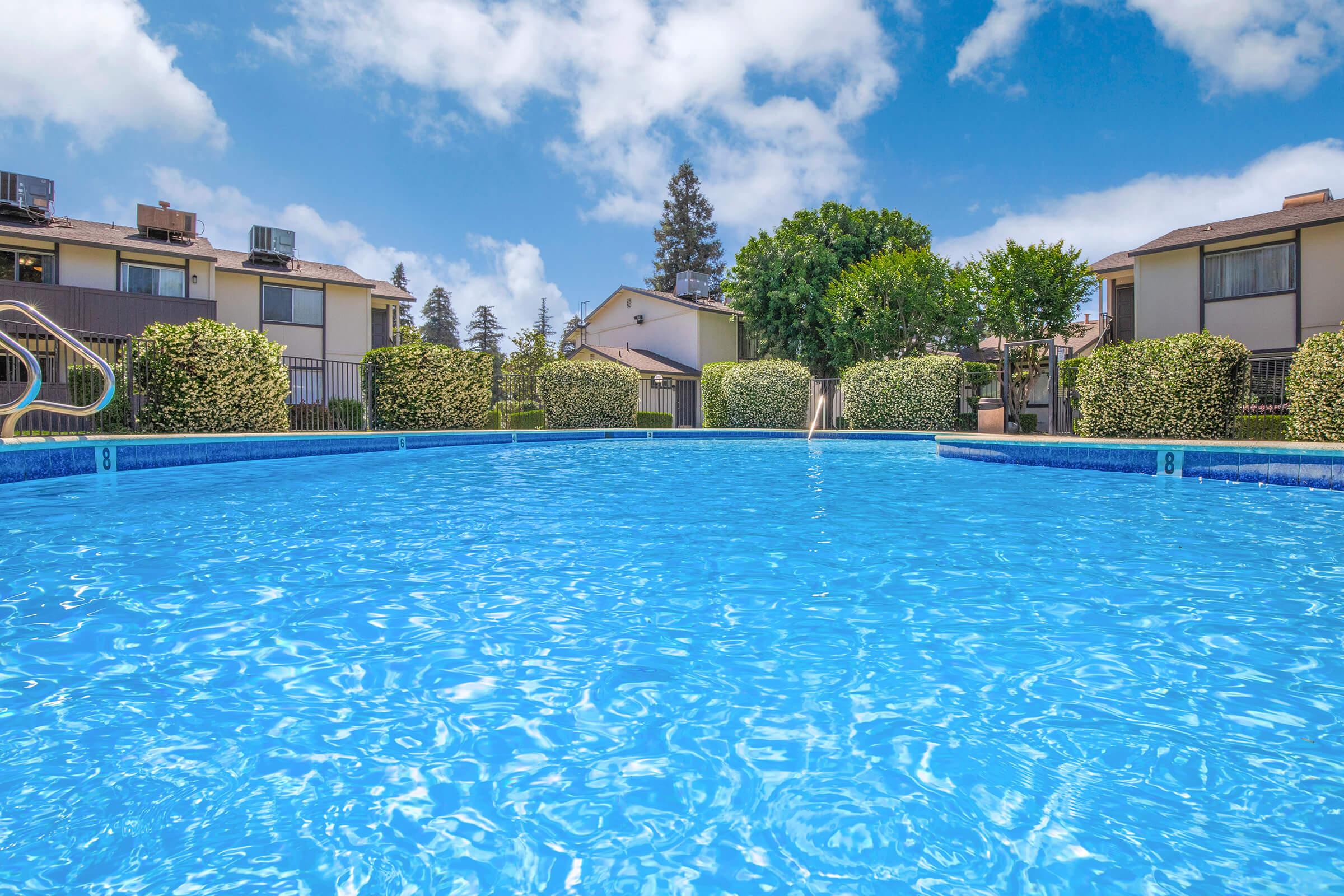 A bright blue swimming pool surrounded by neatly trimmed hedges and trees, with residential buildings in the background under a partly cloudy sky. The water's surface reflects the sunlight, creating a serene and inviting atmosphere.