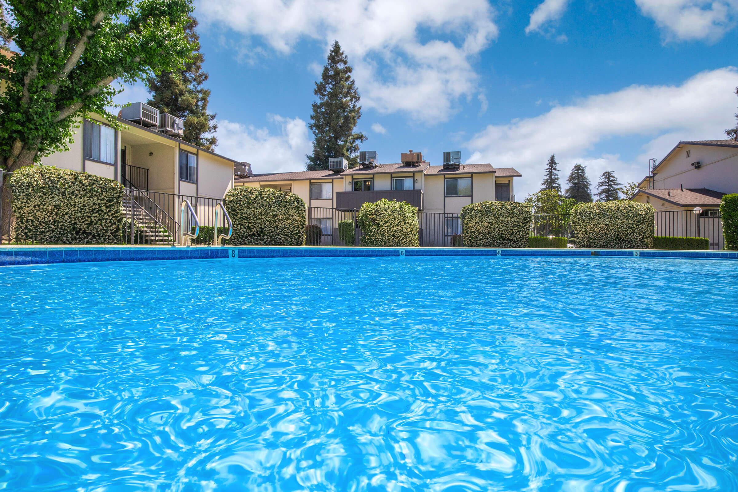Swimming pool with clear blue water in the foreground, surrounded by manicured shrubs and trees. In the background, there are several apartment buildings under a bright blue sky with fluffy white clouds. The setting conveys a serene and inviting atmosphere.