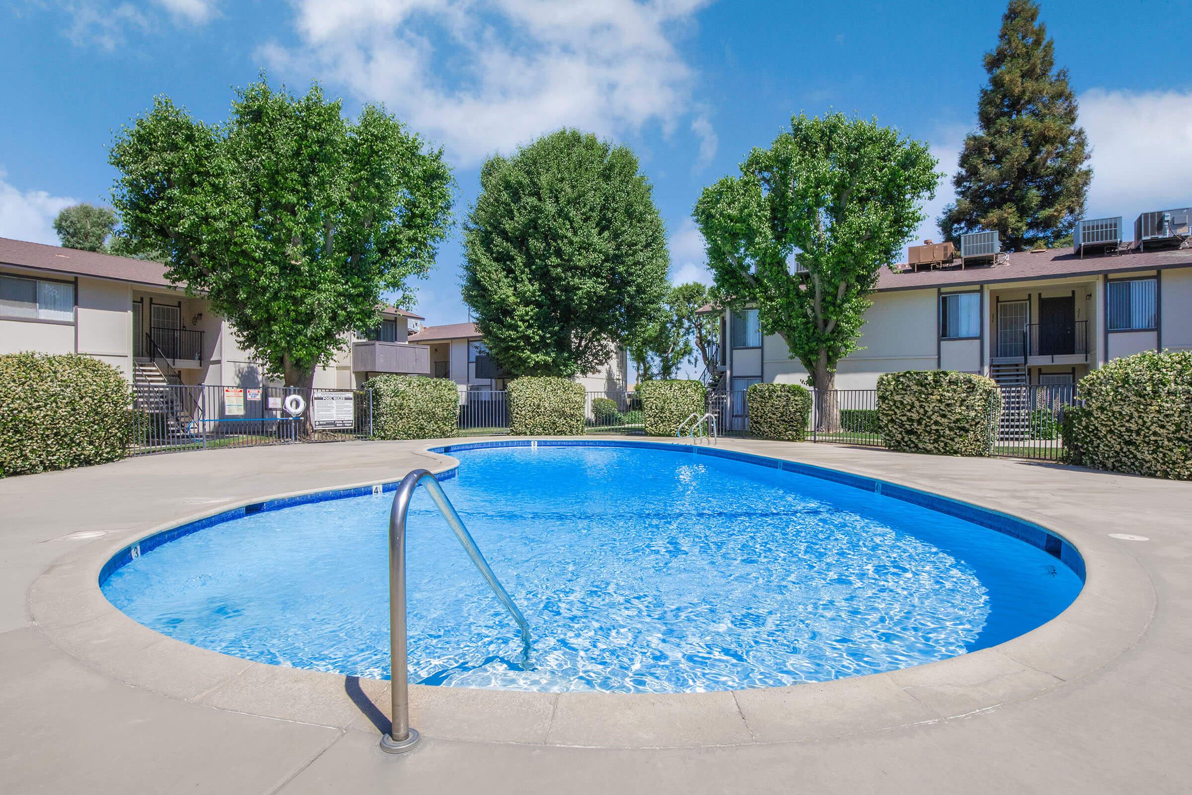 A clear blue swimming pool surrounded by well-manicured lawns and trees. The pool has a gentle slope leading to the water, with steps on one side for entry. In the background, there are two buildings with balconies and windows, set against a bright blue sky with a few clouds.
