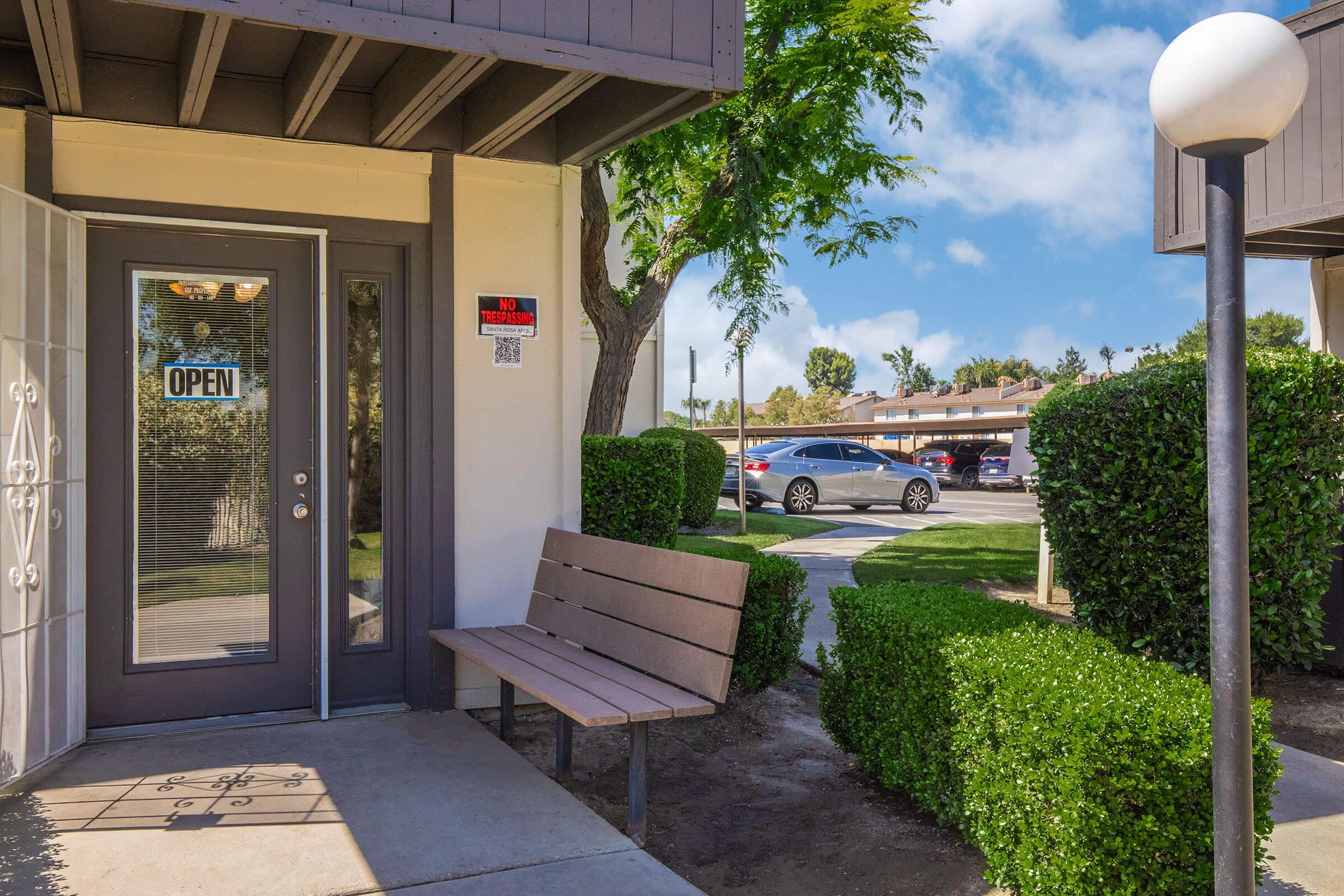 A welcoming entrance featuring a door with an "OPEN" sign, a wooden bench, neatly trimmed bushes, and clear blue skies. In the background, parked cars and additional greenery can be seen, creating a pleasant outdoor atmosphere.
