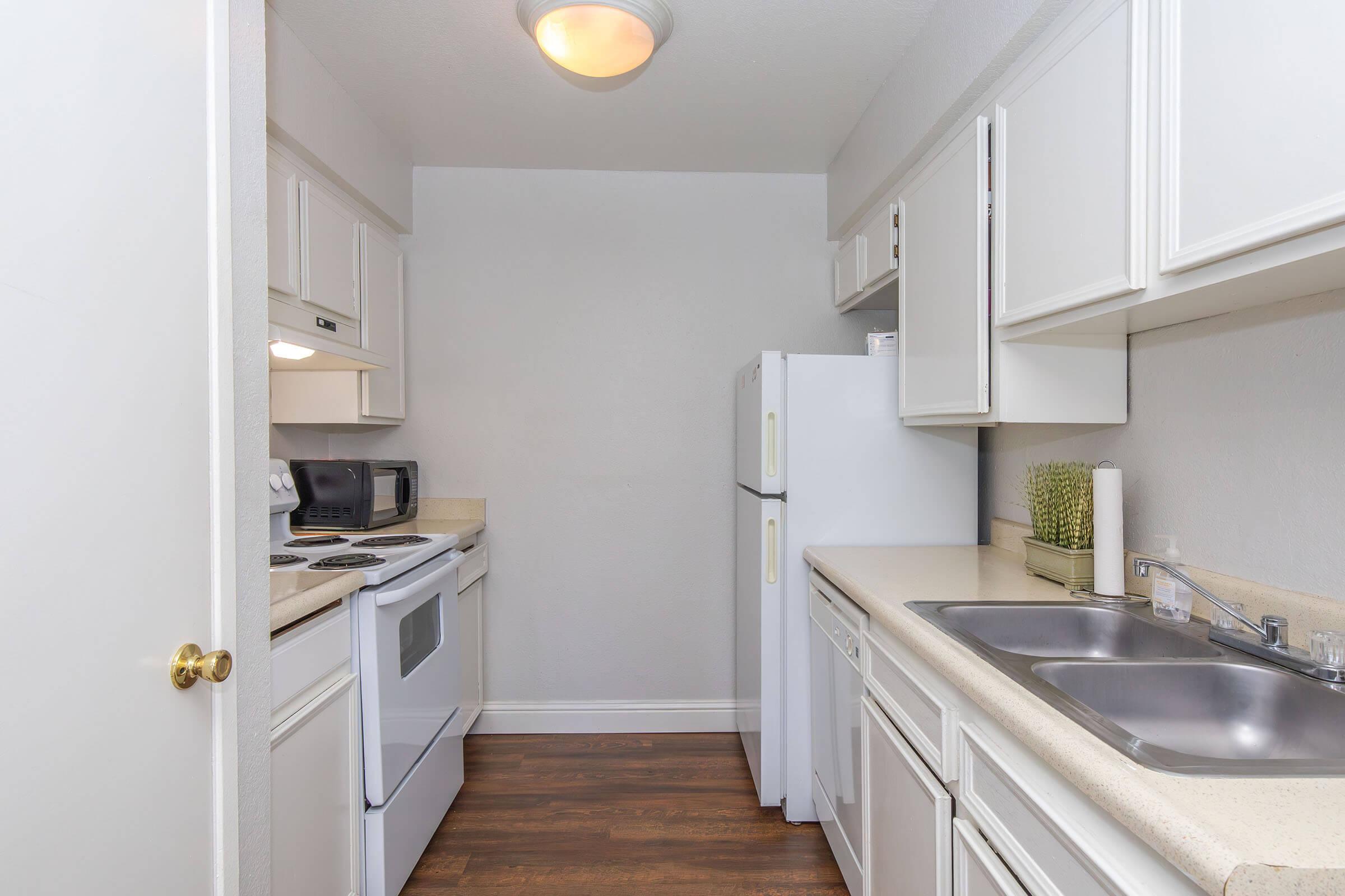 A modern, well-lit kitchen featuring white cabinetry, a double sink, a refrigerator, and an oven with a stovetop. Stainless steel appliances are integrated, including a microwave. The countertops are light-colored, and the flooring is a dark wood, creating a contrasting and inviting space.