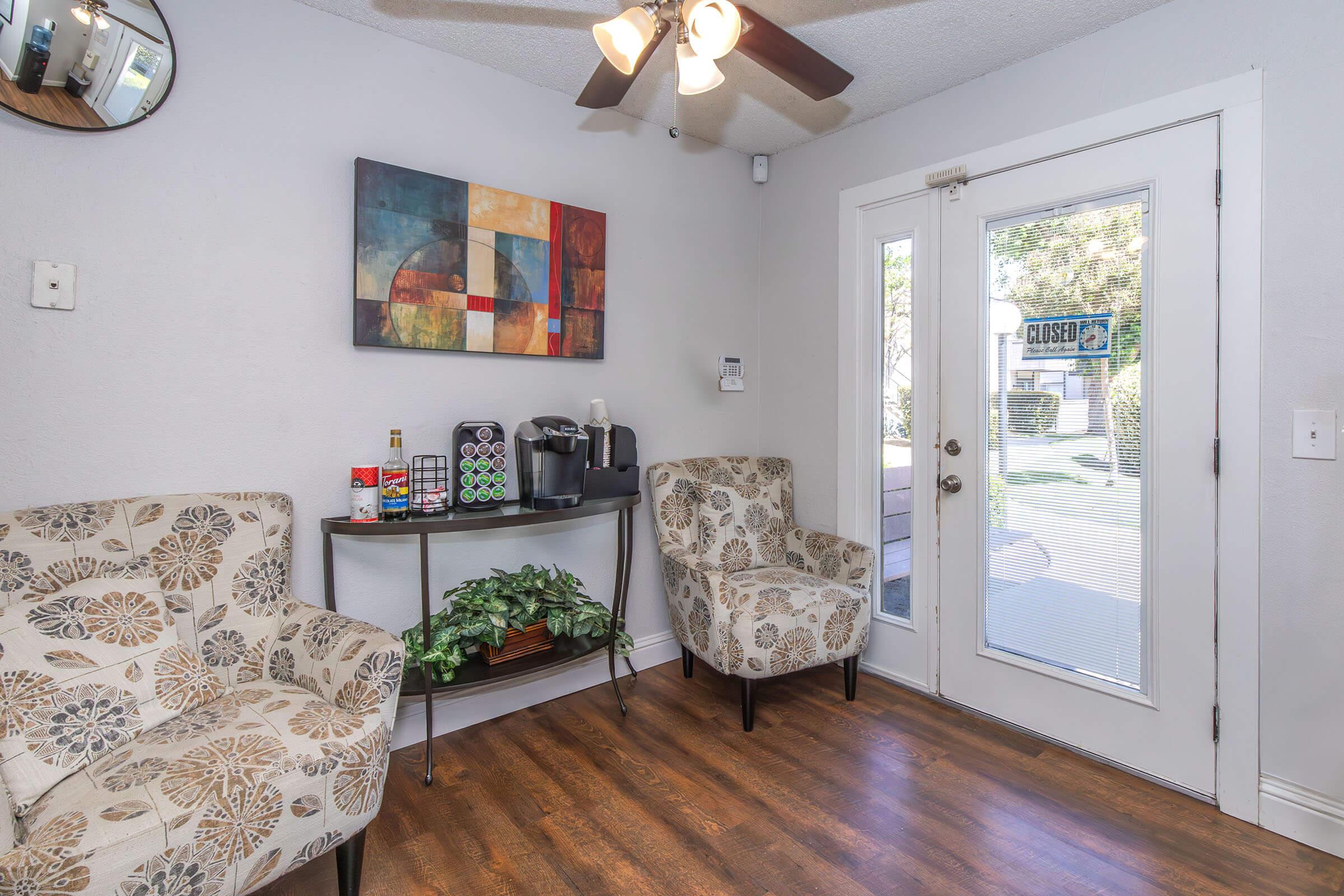 Cozy interior of a small waiting area featuring two patterned armchairs, a decorative table with a coffee machine and beverage options, and a door with a "CLOSED" sign. A round mirror is mounted on the wall, and there are green plants adding a touch of nature to the space.