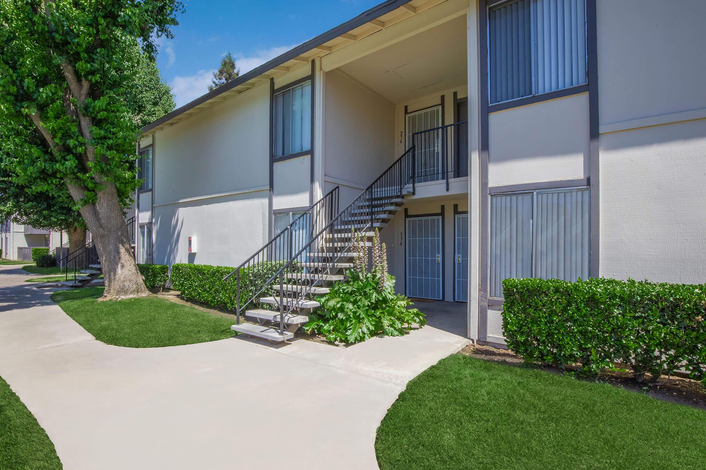 A courtyard scene featuring an apartment building with a staircase leading to the second floor. The exterior has a light color scheme, large windows, and is surrounded by neatly trimmed bushes and grass. A tree is visible to the left, and there is a paved pathway in the foreground.