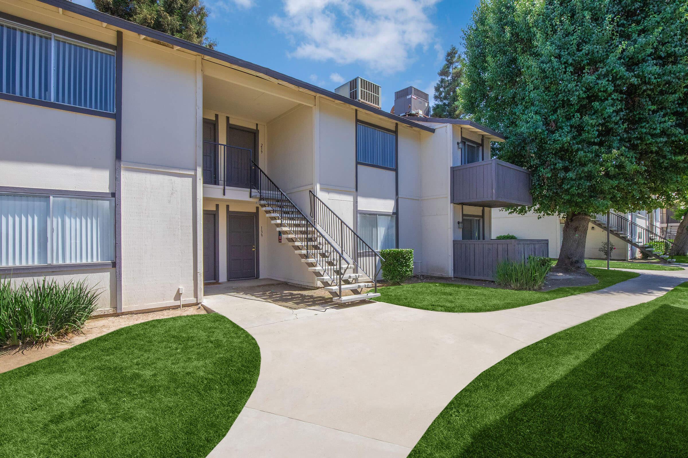 A view of a modern apartment complex featuring a two-story building with a staircase leading to the second floor. Lush green grass surrounds the walkway, and trees are visible in the background. The scene is bright and inviting, with clear blue skies above.