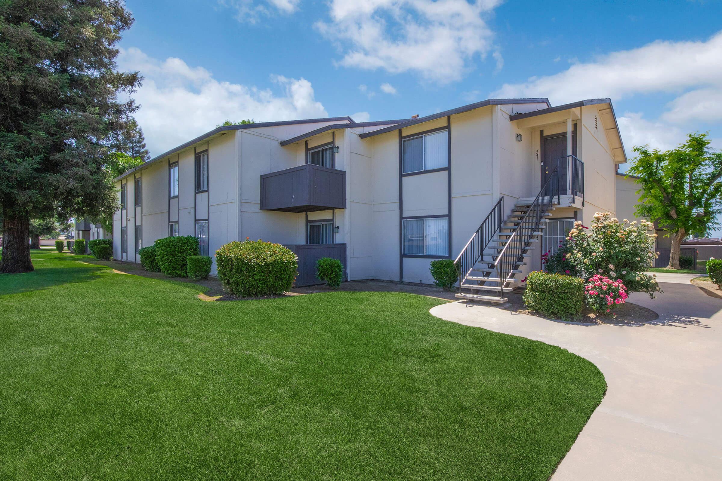Two-story apartment building with a light-colored exterior, surrounded by well-manicured green lawns and neatly trimmed bushes. A staircase leads to the upper floor, and the sky is clear with a few clouds. Pathways connect the building to landscaped areas.