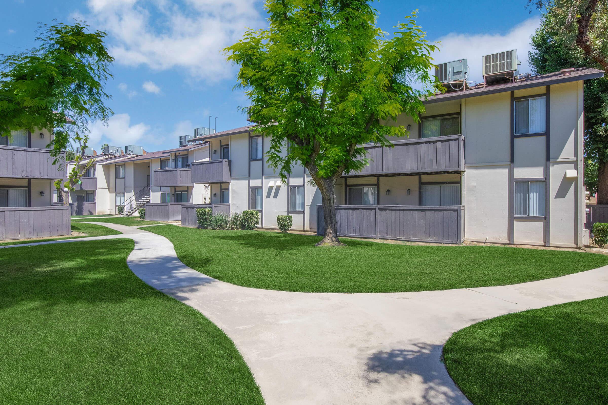 A landscaped apartment complex featuring multiple two-story buildings surrounded by green lawns and trees. A curving walkway connects the buildings, under a clear blue sky with a few clouds.