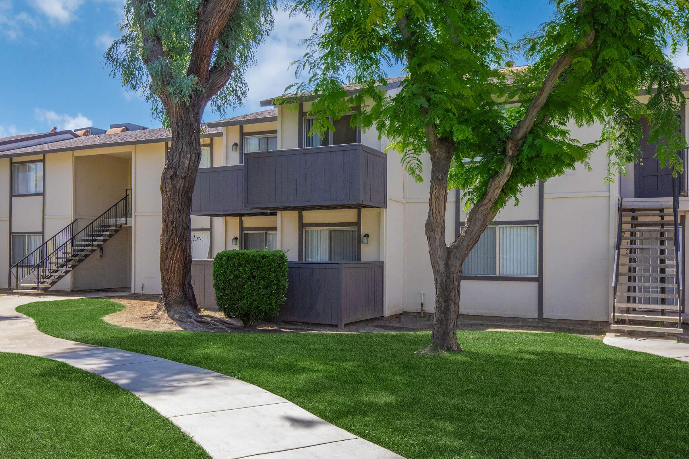 Two-story apartment building with a modern design, featuring balconies and large windows. Green grass and a curved pathway in the foreground, bordered by trees. The sky is clear with a few clouds, creating a bright and inviting atmosphere.