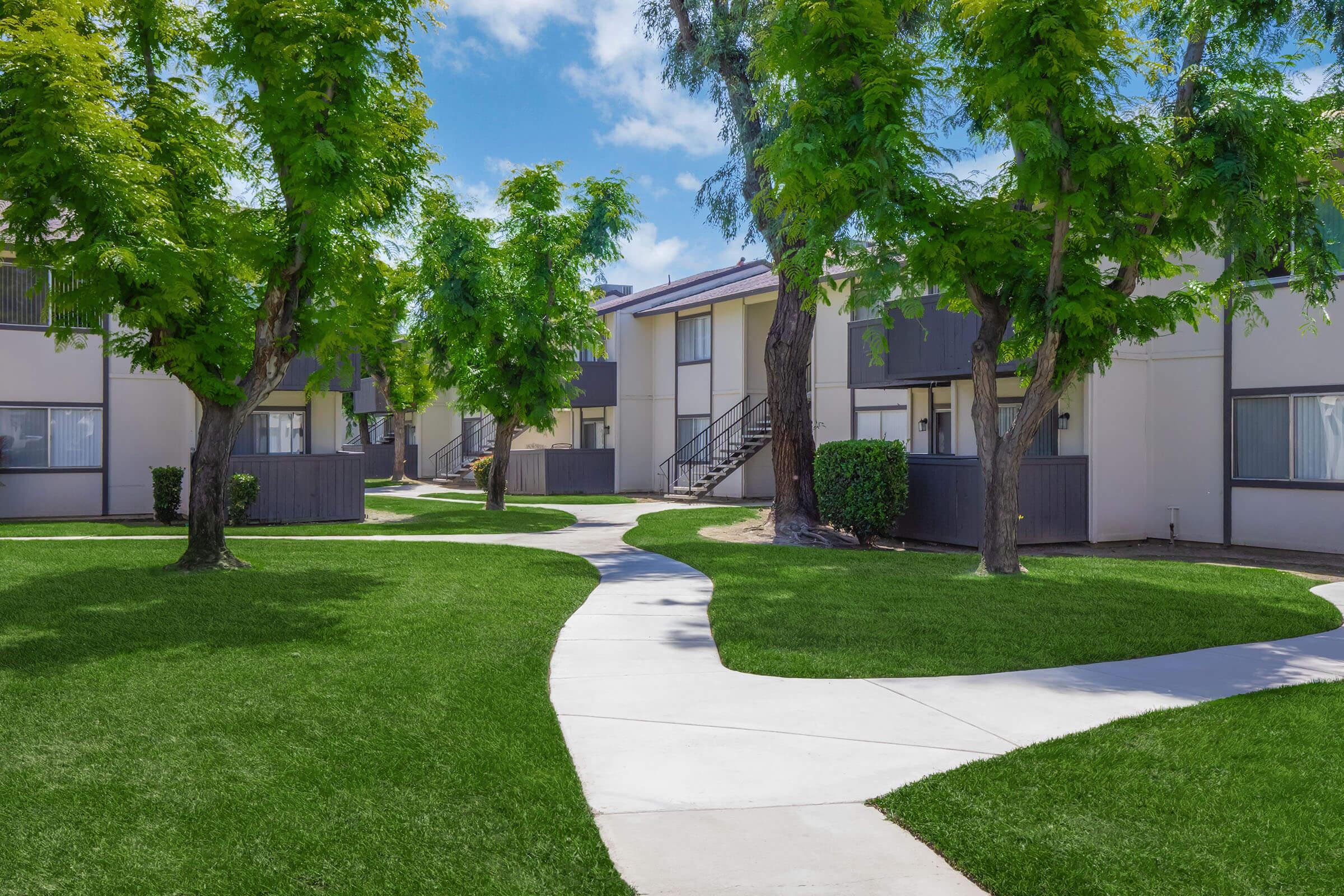 A landscaped apartment complex featuring well-maintained green grass, trees, and a paved walkway. The buildings are low-rise with a modern design, providing a serene outdoor environment suitable for residents. The sky is clear with a few clouds, adding to the pleasant atmosphere.