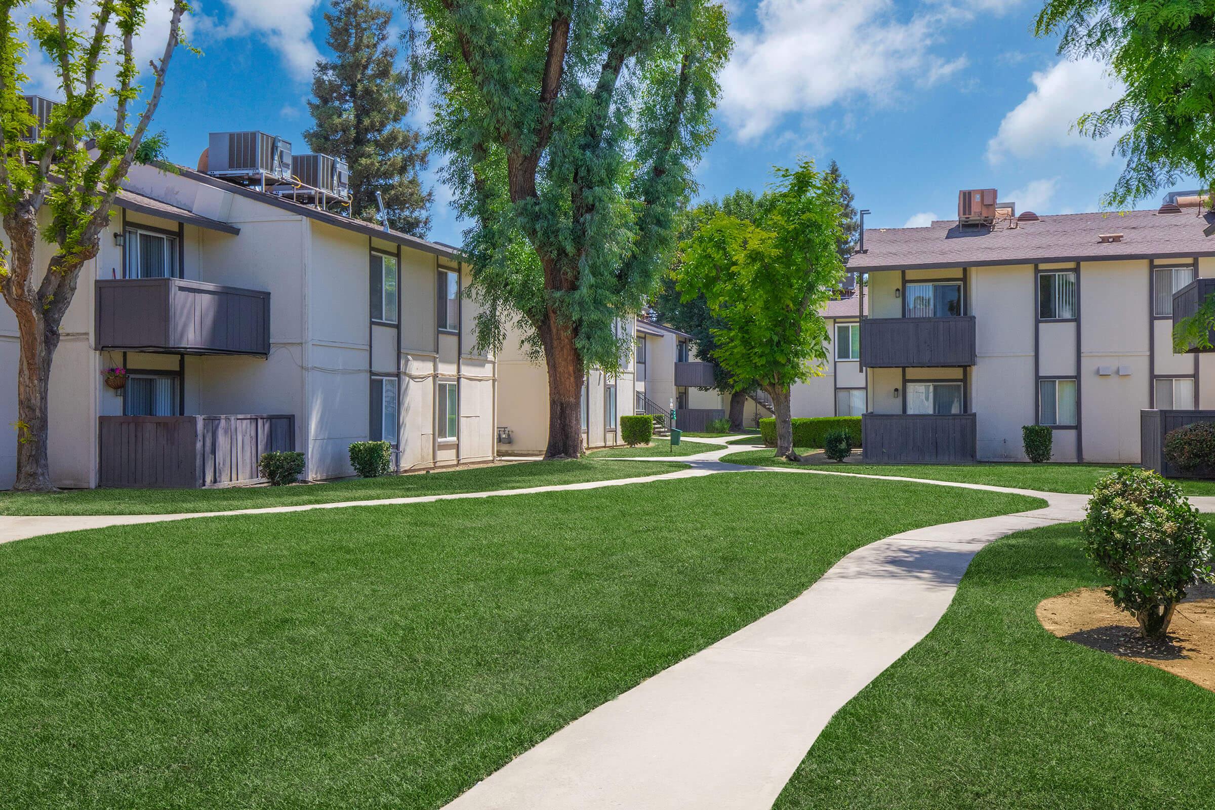 A landscaped courtyard featuring two apartment buildings, surrounded by vibrant green grass. The pathway winds through the lawn, connecting the buildings and bordered by shrubs and trees, under a clear blue sky with a few clouds.