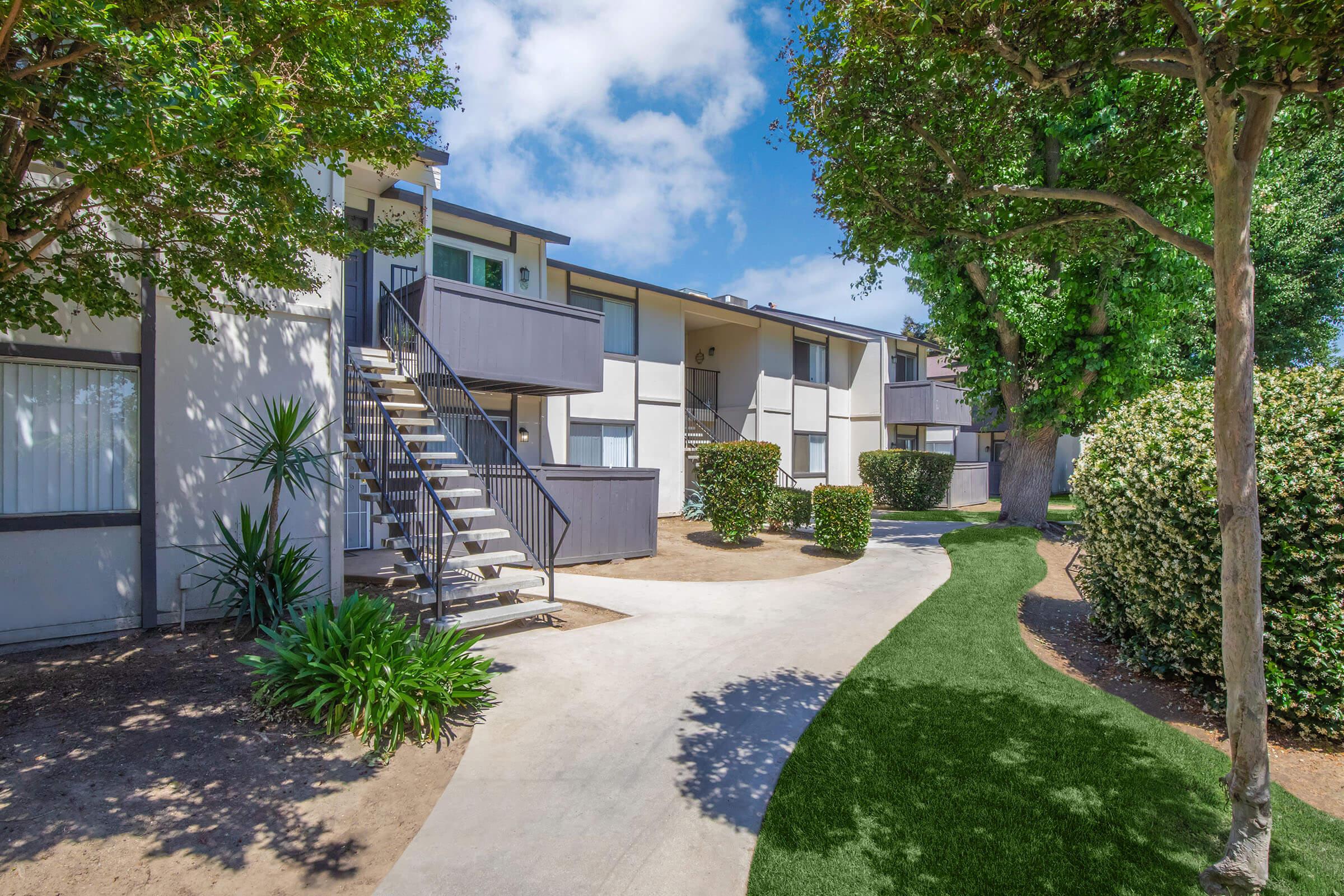 A modern apartment complex featuring two stories with exterior staircases, surrounded by neatly trimmed hedges and green lawns. The scene includes landscaped pathways and trees against a bright blue sky with scattered clouds.