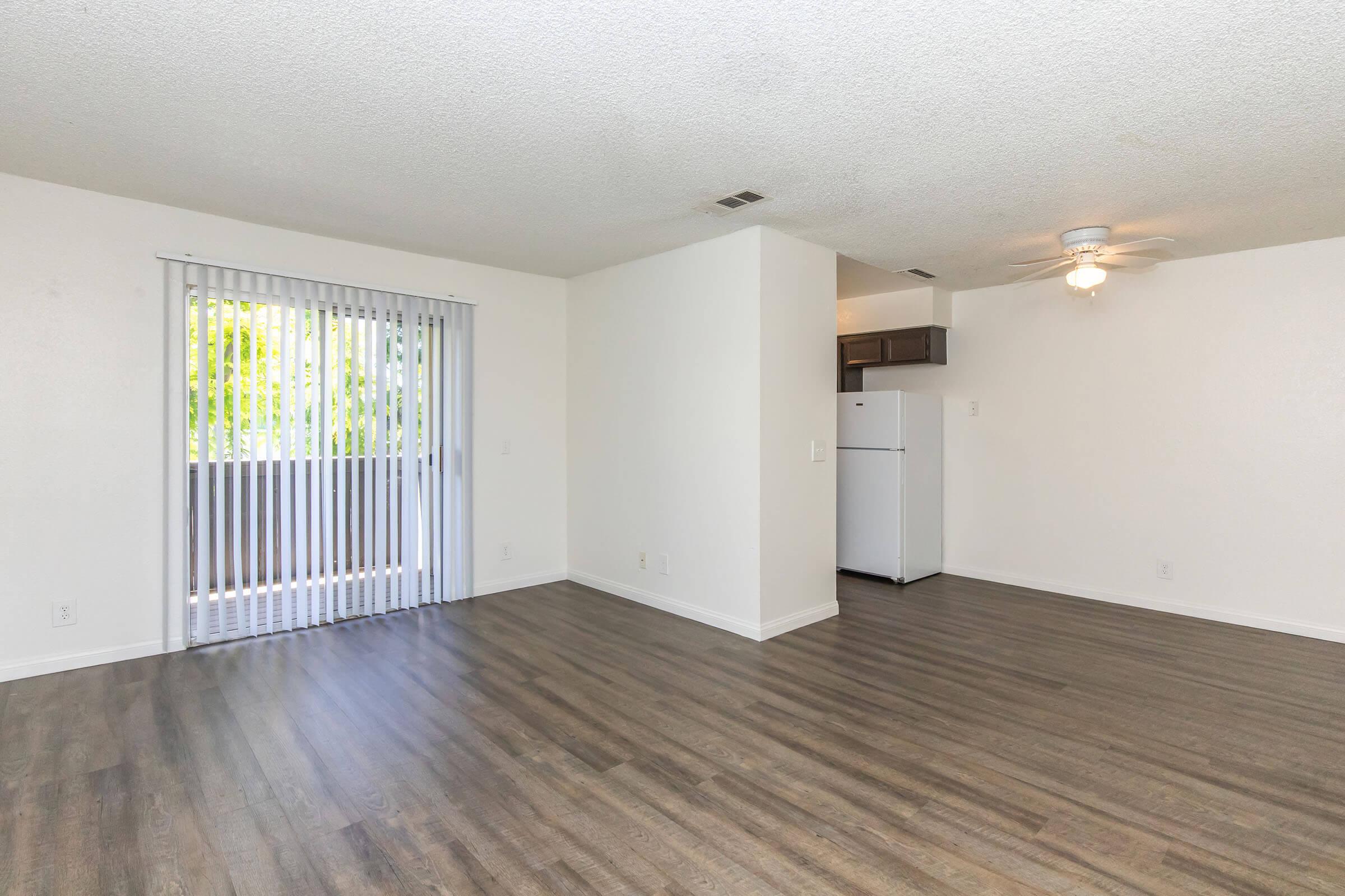 Bright and spacious living area with laminate flooring, featuring a sliding glass door leading to a balcony. A ceiling fan is visible, along with a white refrigerator in the kitchen area. The walls are painted light colors, providing an inviting atmosphere.