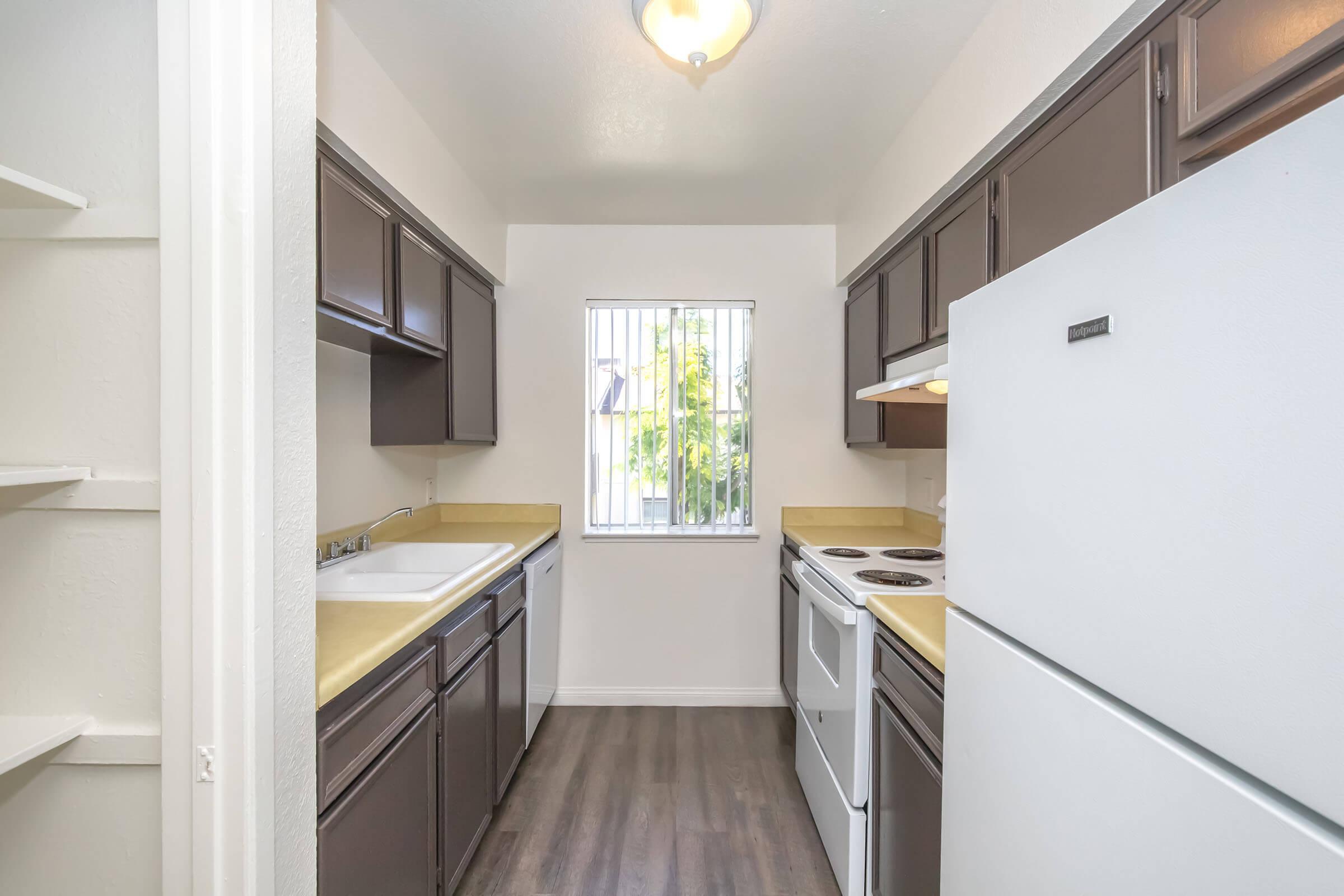 A narrow kitchen with dark wooden cabinets, a white refrigerator, an oven, and a stovetop. There's a window with blinds allowing natural light, and a small shelf on the left. The countertops are light-colored, and the floor has a wood-like finish. Overall, the space appears clean and organized.