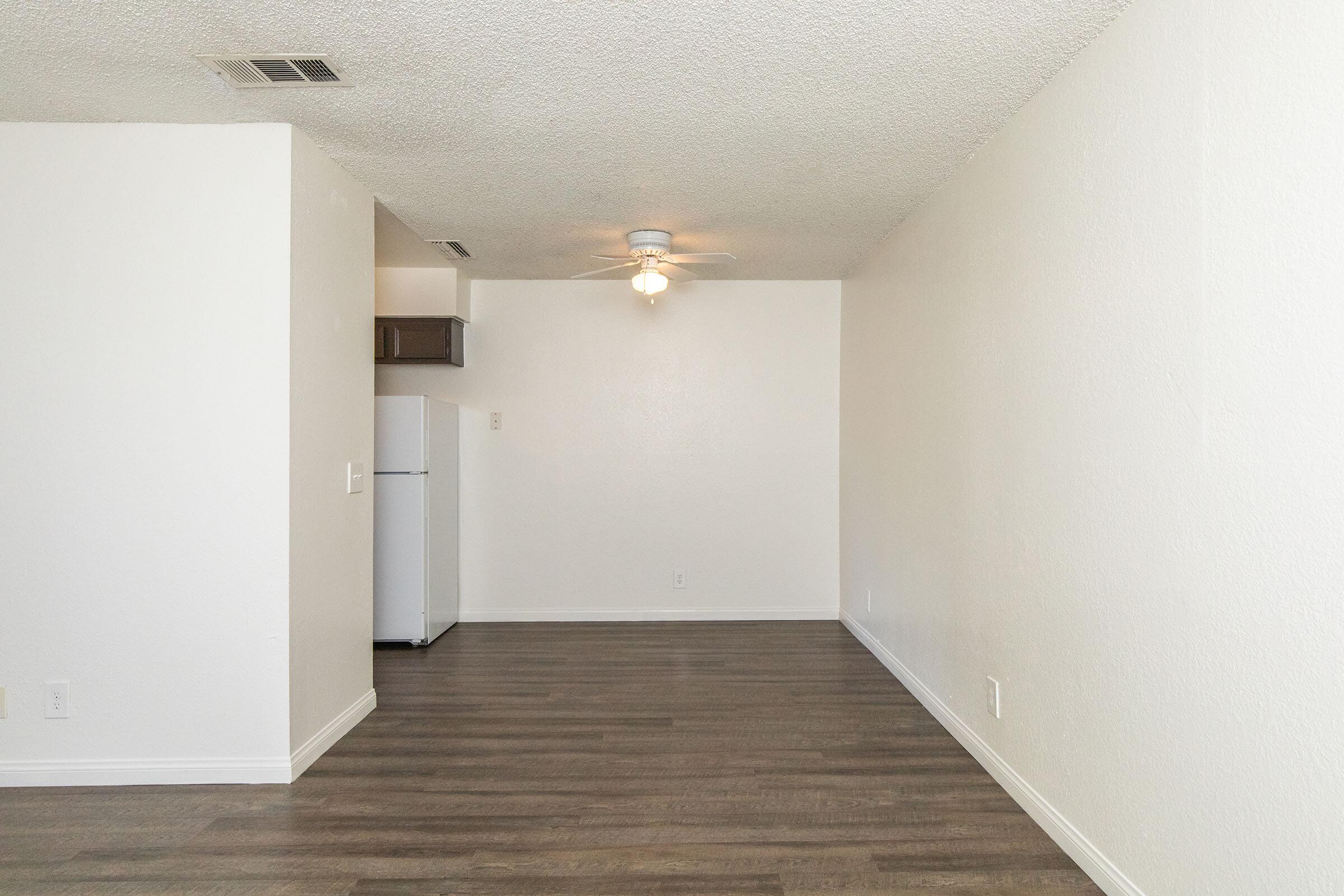 Spacious room with light-colored walls and a ceiling fan, featuring laminate flooring. A small kitchenette is visible in the background, showcasing a white refrigerator and minimal cabinetry. The space is well-lit and designed for a cozy living area.