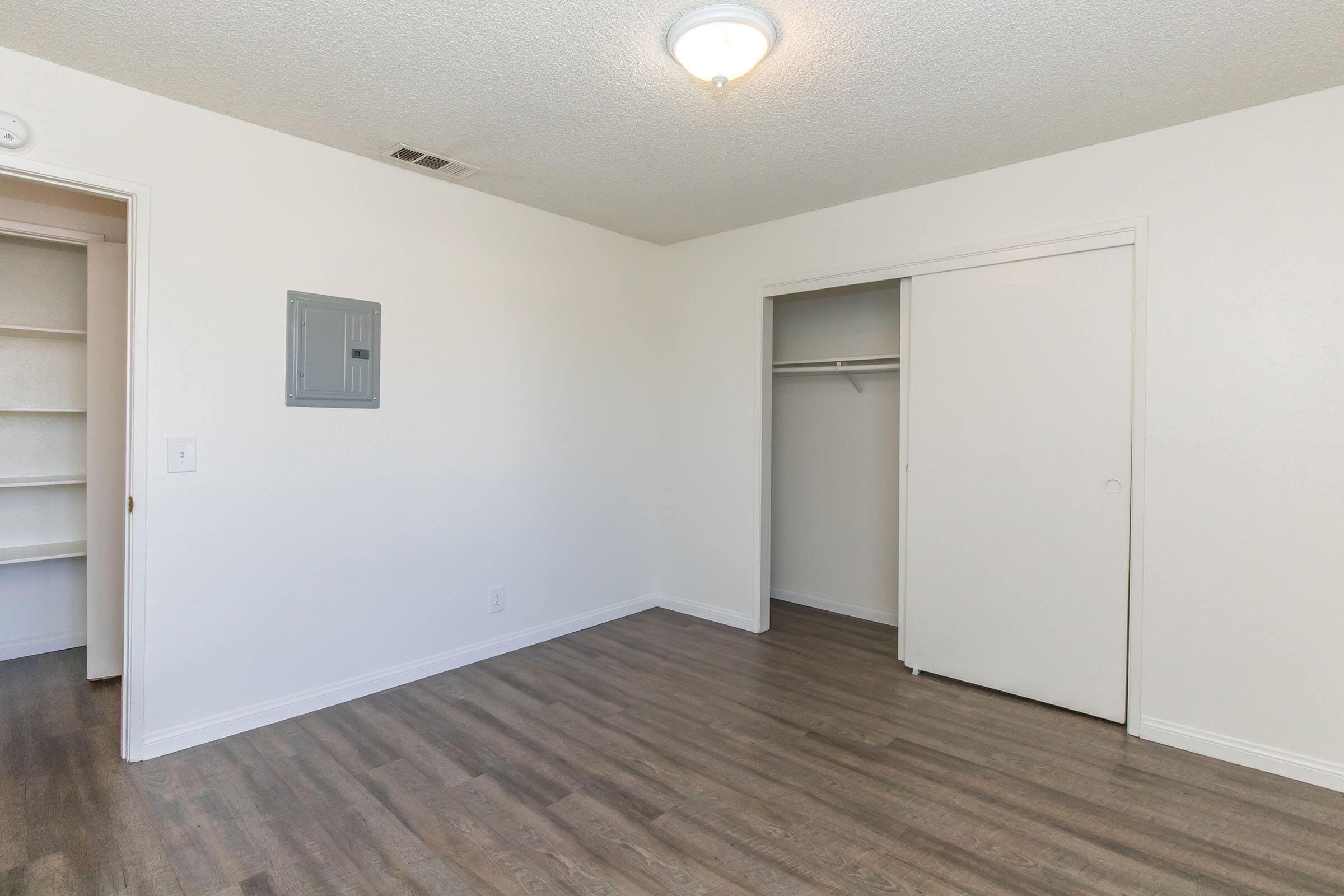 A spacious, empty room with light-colored walls and wood-style flooring. There is a closet with sliding doors on the right and a small utility box on the left wall. A ceiling light illuminates the area, and the overall feel is bright and airy.