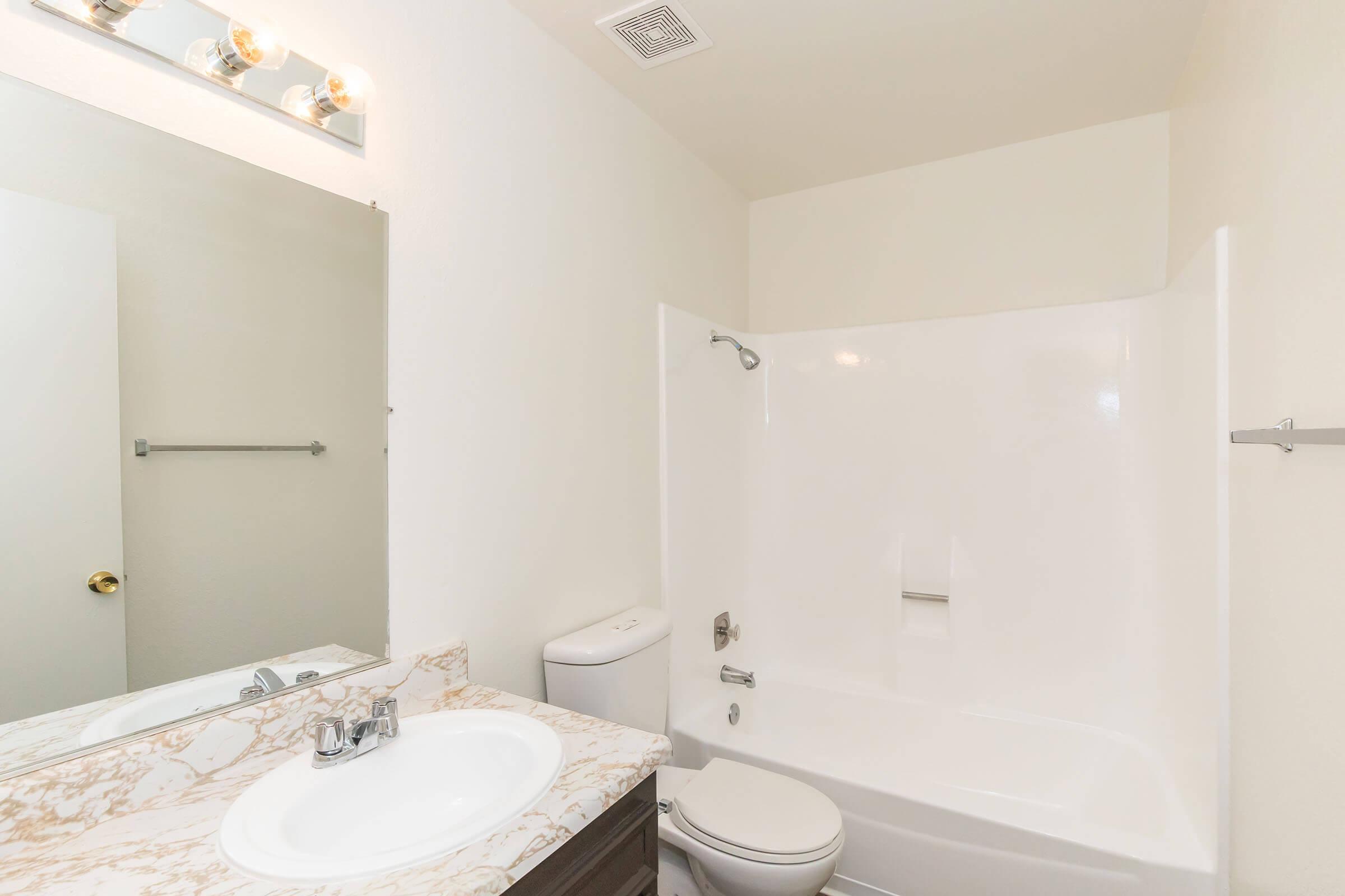 A clean and modern bathroom featuring a white tub and shower, a sink with a marble countertop, a mirror above the sink, and a toilet. The walls are painted white, and there is a small towel bar next to the mirror. The overall design is simple and functional.