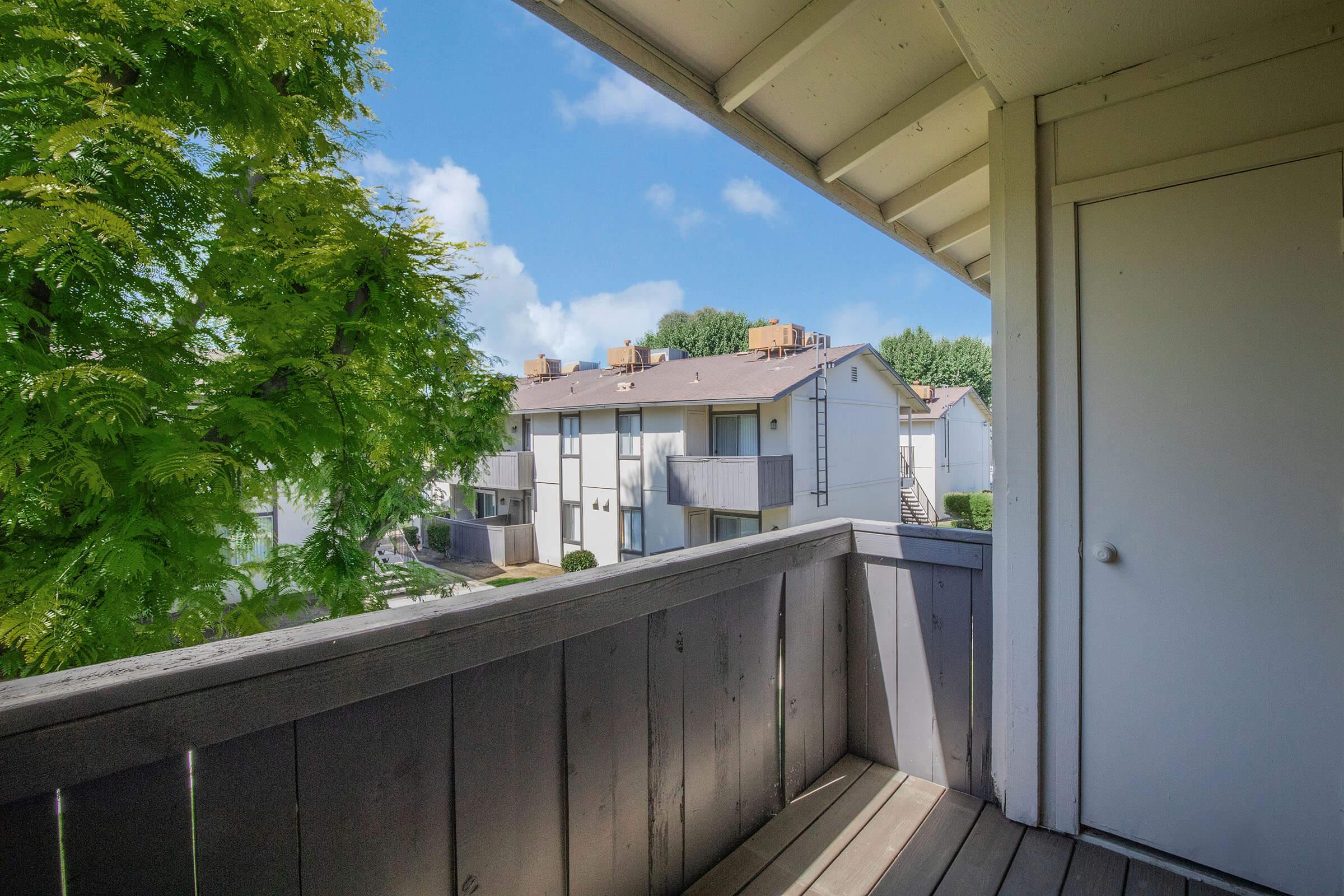 A view from a balcony showing a tree and nearby apartment buildings under a clear blue sky. The scene captures a peaceful residential area, emphasizing greenery and a well-maintained environment. The balcony features a wooden railing and a door leading into the interior space.