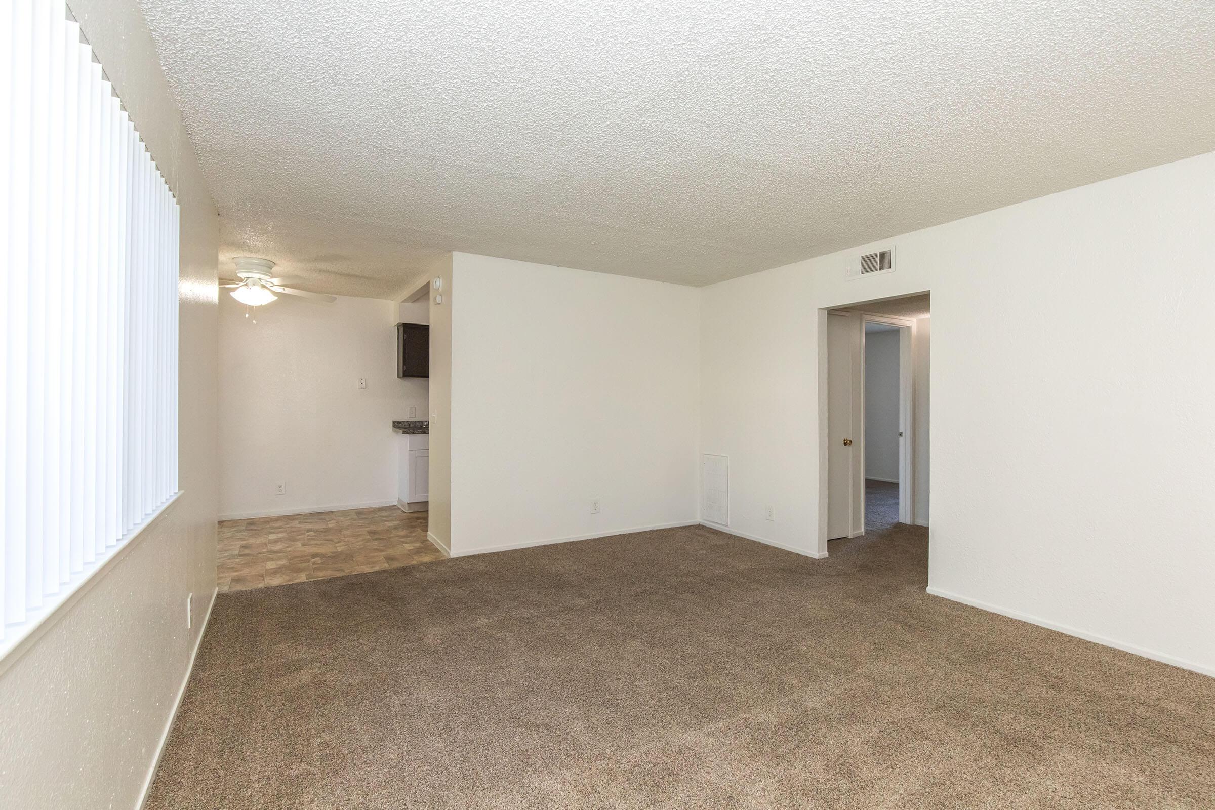A spacious, empty living room with beige carpet and white textured walls. A ceiling fan is visible in the upper left corner. To the back left, there is a doorway leading to a kitchen area with tiled flooring. A second doorway on the right leads to another room, creating an open and well-lit space.
