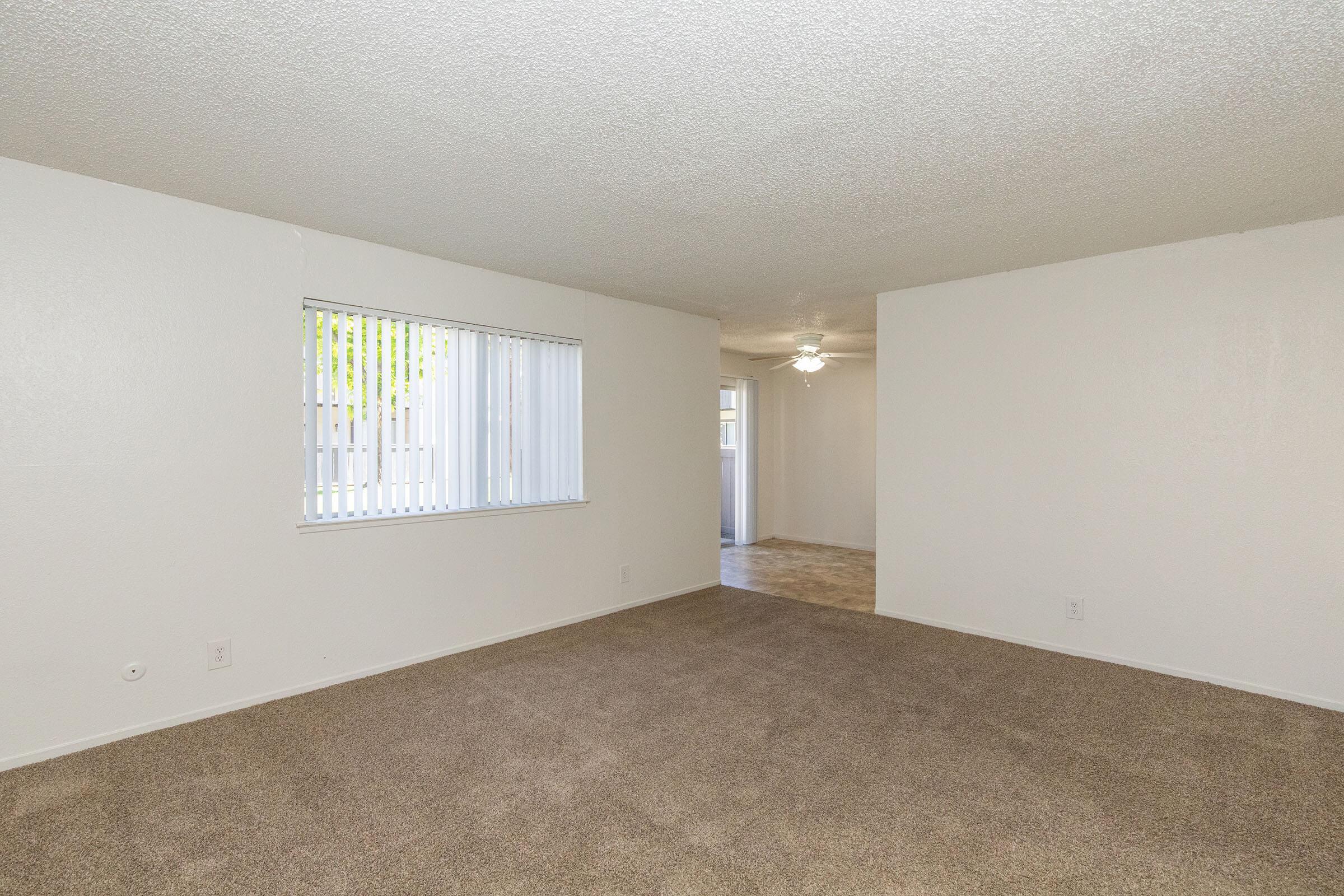 A spacious, empty living room featuring beige carpet and white walls. A window with vertical blinds lets in natural light, and a ceiling fan is visible in the distance. The room connects to another area, suggesting an open layout.