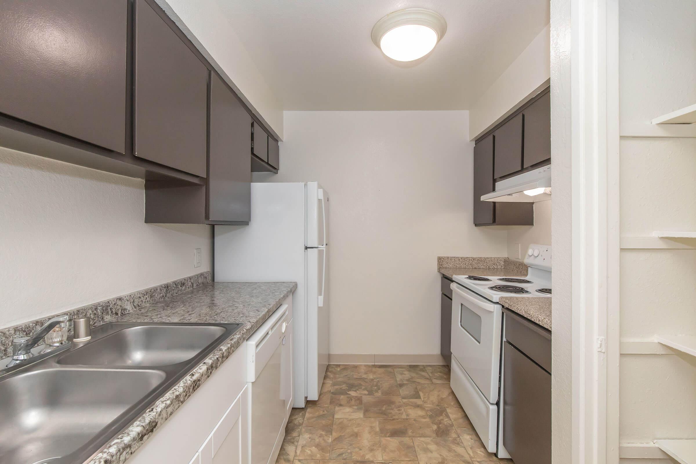 A clean and modern kitchen featuring dark cabinets, a white stove and refrigerator, and a double sink with a granite countertop. The kitchen has a neutral wall color and a light fixture above, with laminate flooring adding to the contemporary feel.