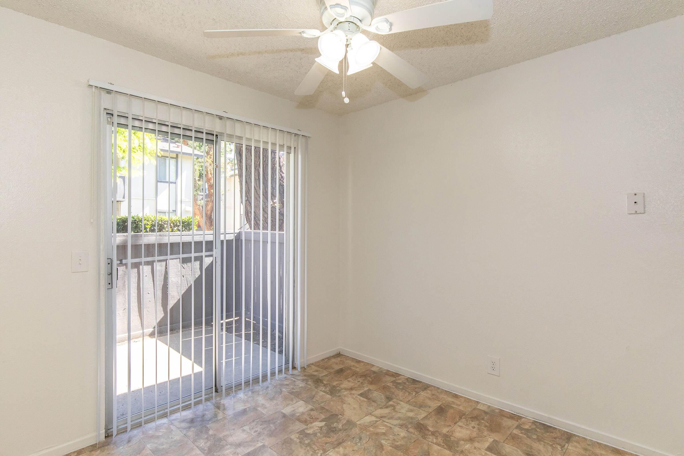 Interior of a room with a ceiling fan, light-colored walls, and a sliding glass door with vertical blinds. The floor features a patterned tile design. Natural light is coming through the door, leading to a small outdoor area.