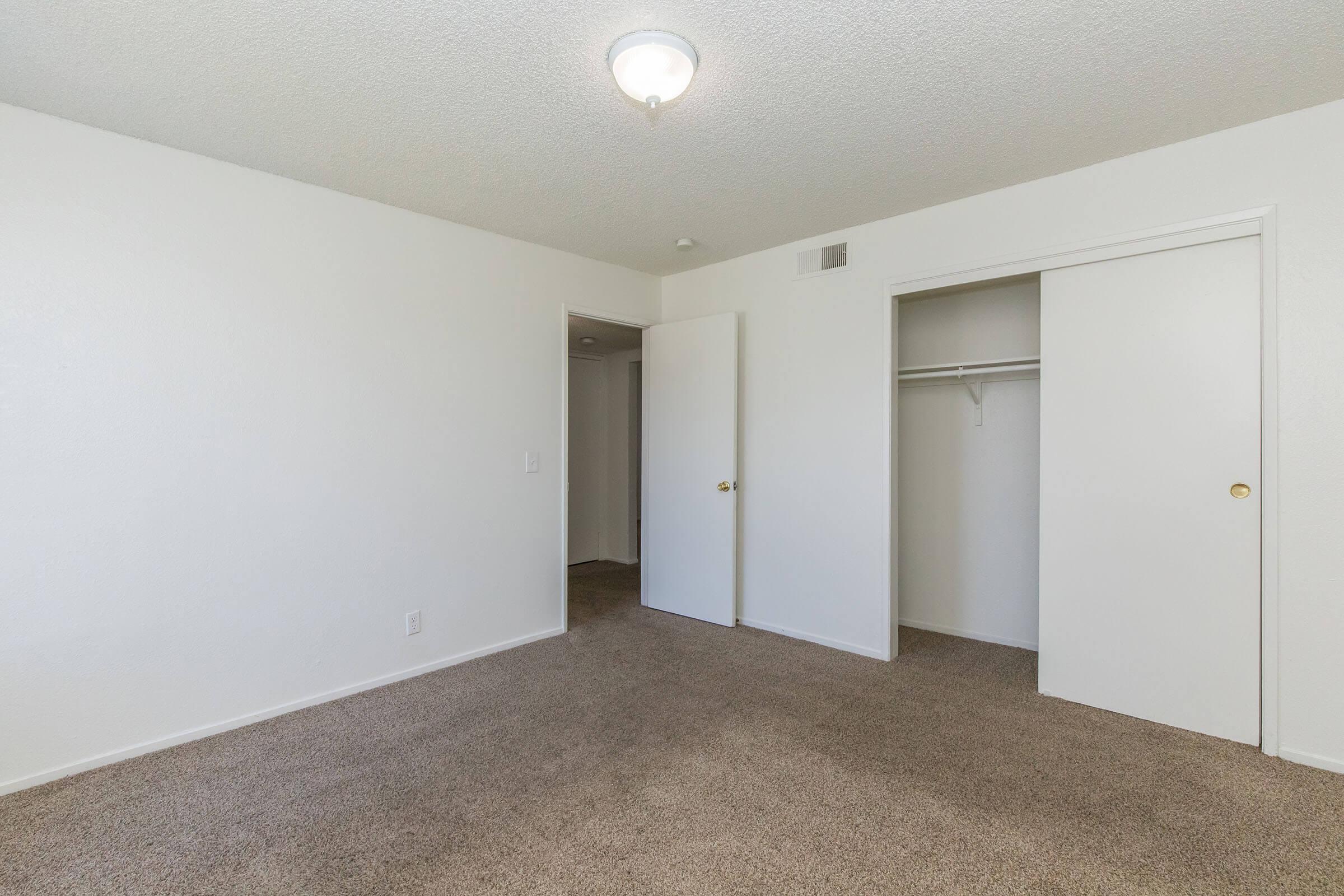 A spacious, empty room with white walls and light brown carpet. There are two doors: one leading to a closet and another possibly to a hallway. A ceiling light fixture is visible, providing overhead illumination. The room appears clean and ready for furnishings or personalization.