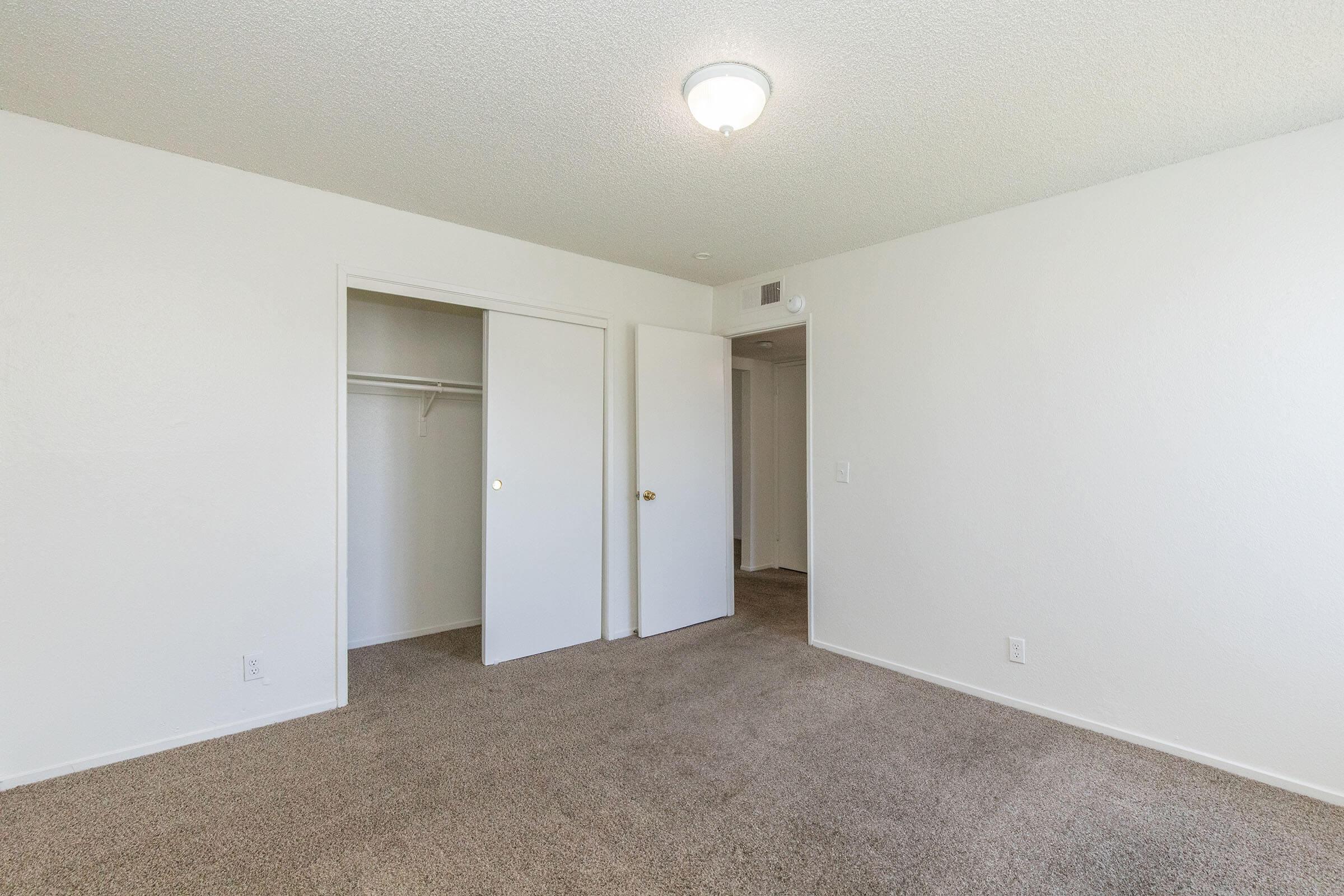 A spacious, empty room with light-colored walls and carpeted floor. There is a ceiling light fixture and two doors: one leading to a closet and another potentially to a hallway. The room has a clean and bright aesthetic, ideal for personalization.