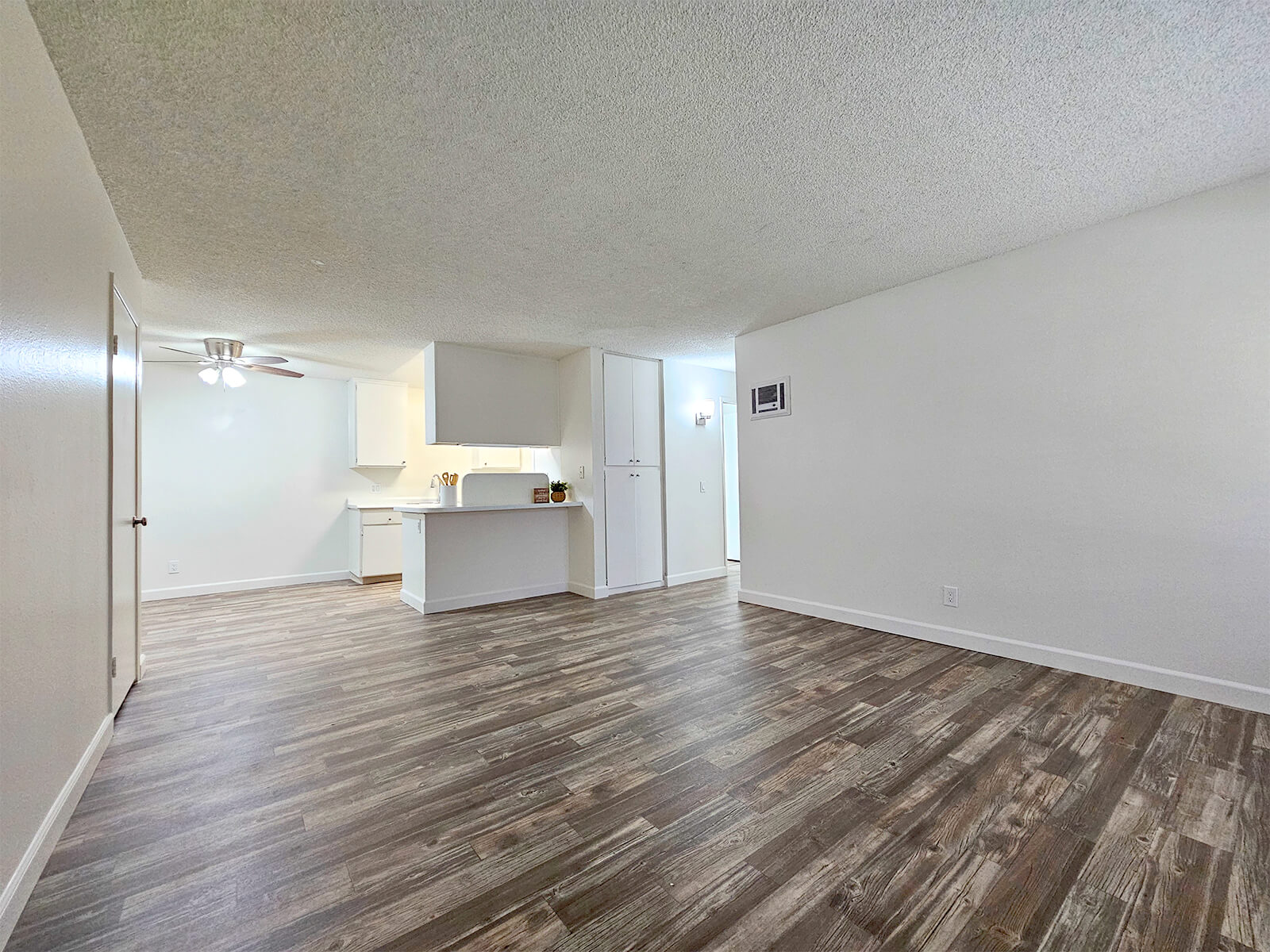 A spacious, empty living area with a wood-like laminate floor. A kitchen is visible in the background, featuring white cabinets and appliances. The ceiling has a fan, and the walls are painted in a light color. Natural light fills the room, creating a bright and airy atmosphere.