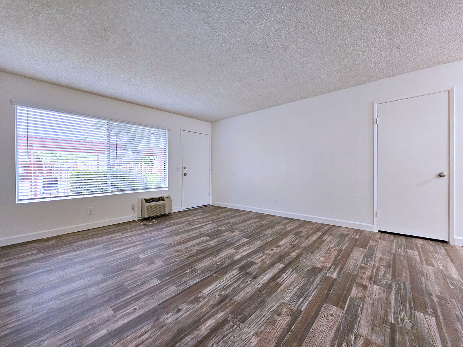 A spacious, well-lit living room featuring a large window with blinds, a door leading outside, and a simple interior design. The flooring is made of wooden planks, and the walls are painted white, creating a clean and modern aesthetic. An air conditioning unit is visible near the window.