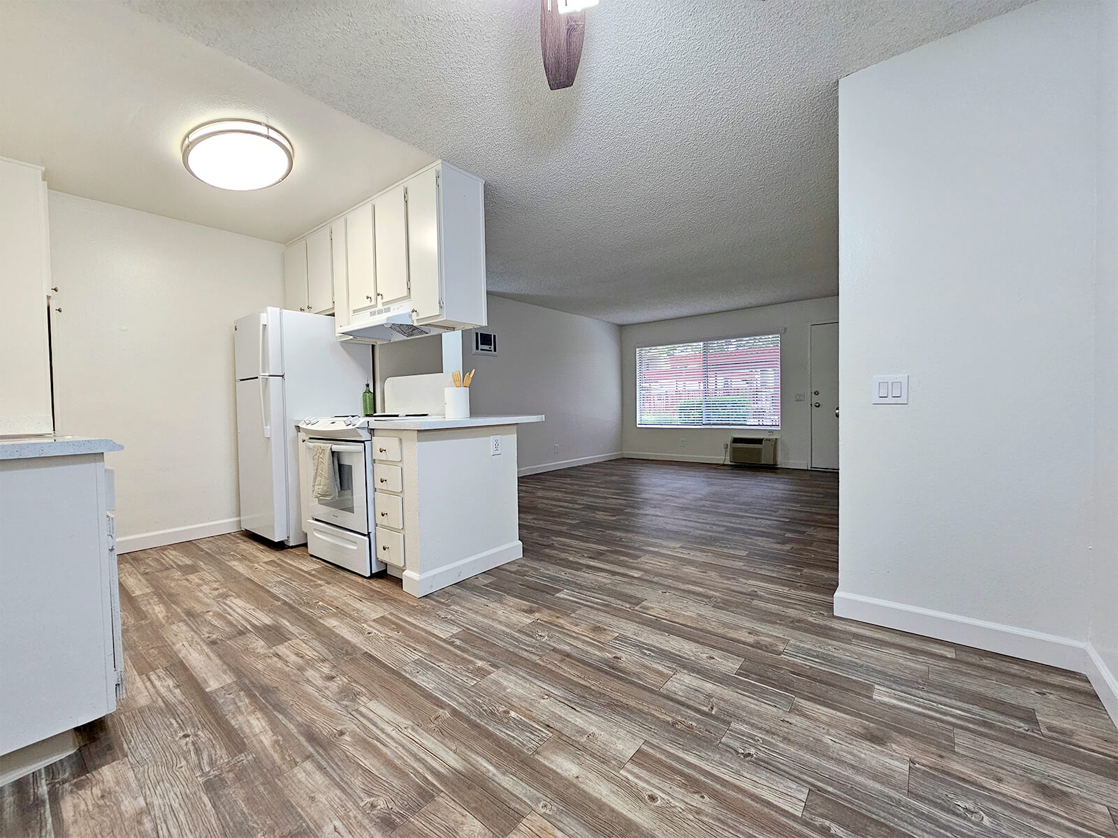 A well-lit apartment interior featuring a kitchen with white cabinets, a refrigerator, and a stove. The open living area has hardwood-style flooring, a large window providing natural light, and a minimalist design.
