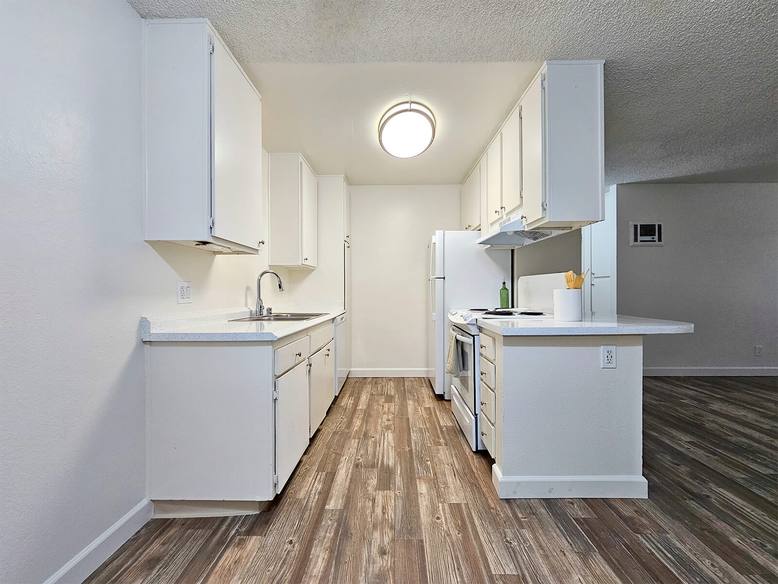 A modern kitchen featuring white cabinets, a stainless steel sink, and white appliances. Natural wood-look flooring complements the design. The room is well-lit with a ceiling light, and there is an open layout leading to another living space.