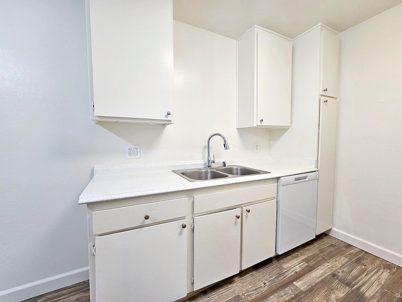 A clean, modern kitchen with white cabinets and a light countertop. It features a double sink with a polished chrome faucet, a dishwasher, and minimalistic open space. The walls are painted white, and the flooring is light wood, creating a bright and inviting atmosphere.