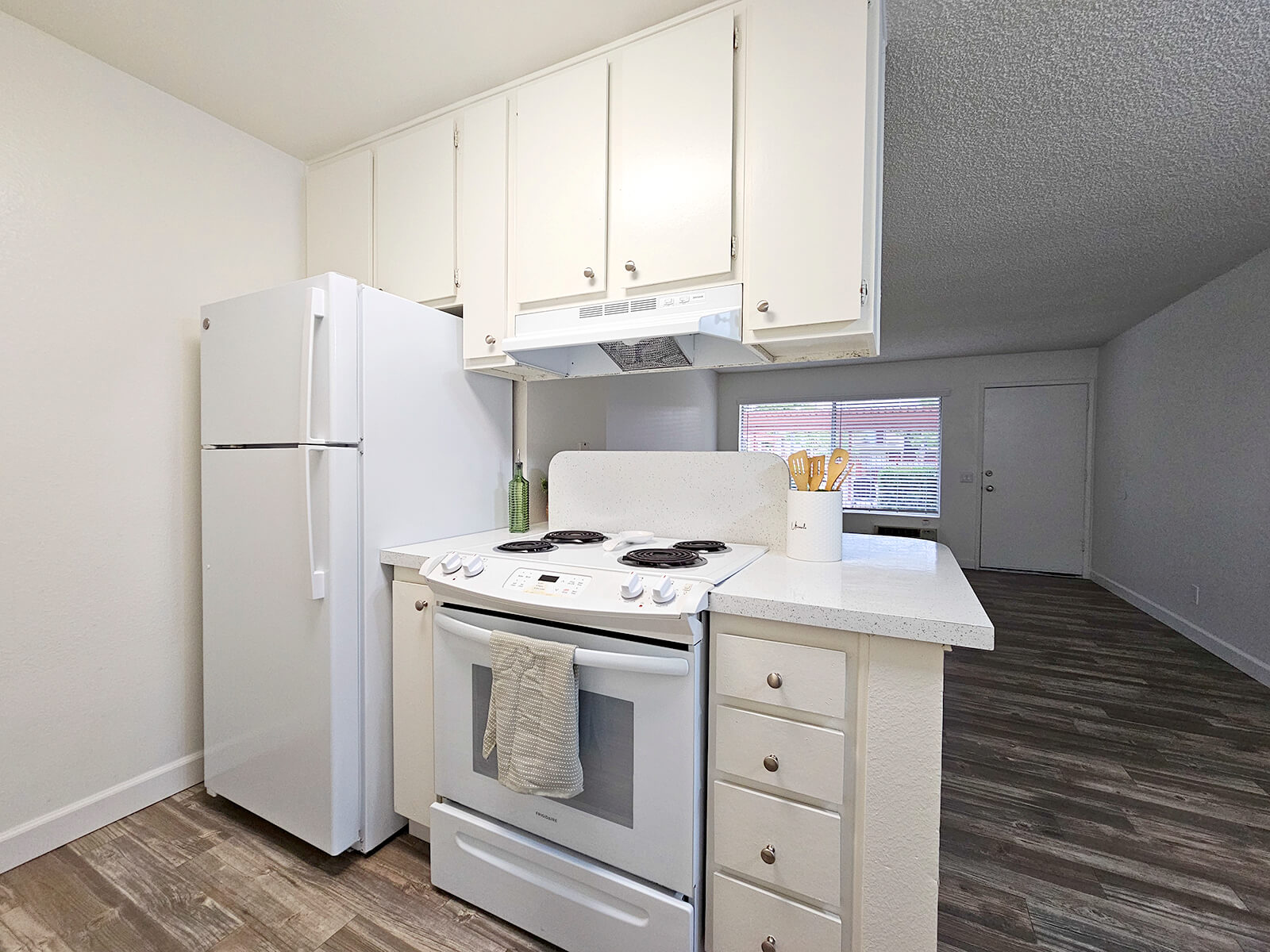 Modern kitchen with white cabinetry, a white refrigerator, and an oven with a stovetop. The countertop is light-colored and features cooking utensils. Natural light filters in through a nearby window, illuminating the space. The flooring is textured and has a wood-like appearance, leading to an open area with a door in the background.