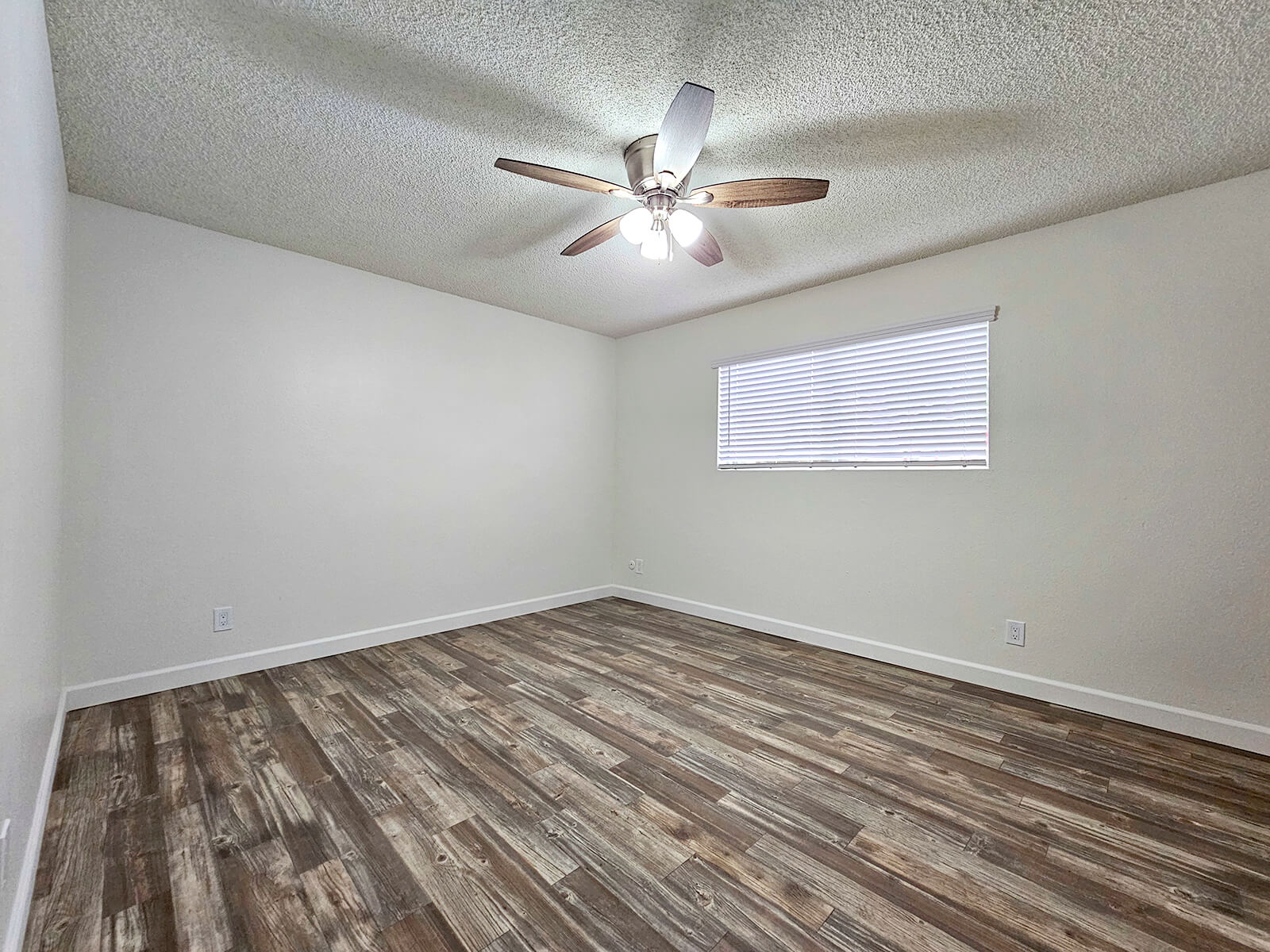 A spacious, empty room featuring a ceiling fan with wooden blades, a light fixture, and a single window with horizontal blinds. The floor has plank-style laminate, and the walls are painted in a light, neutral color, creating a bright and airy atmosphere.