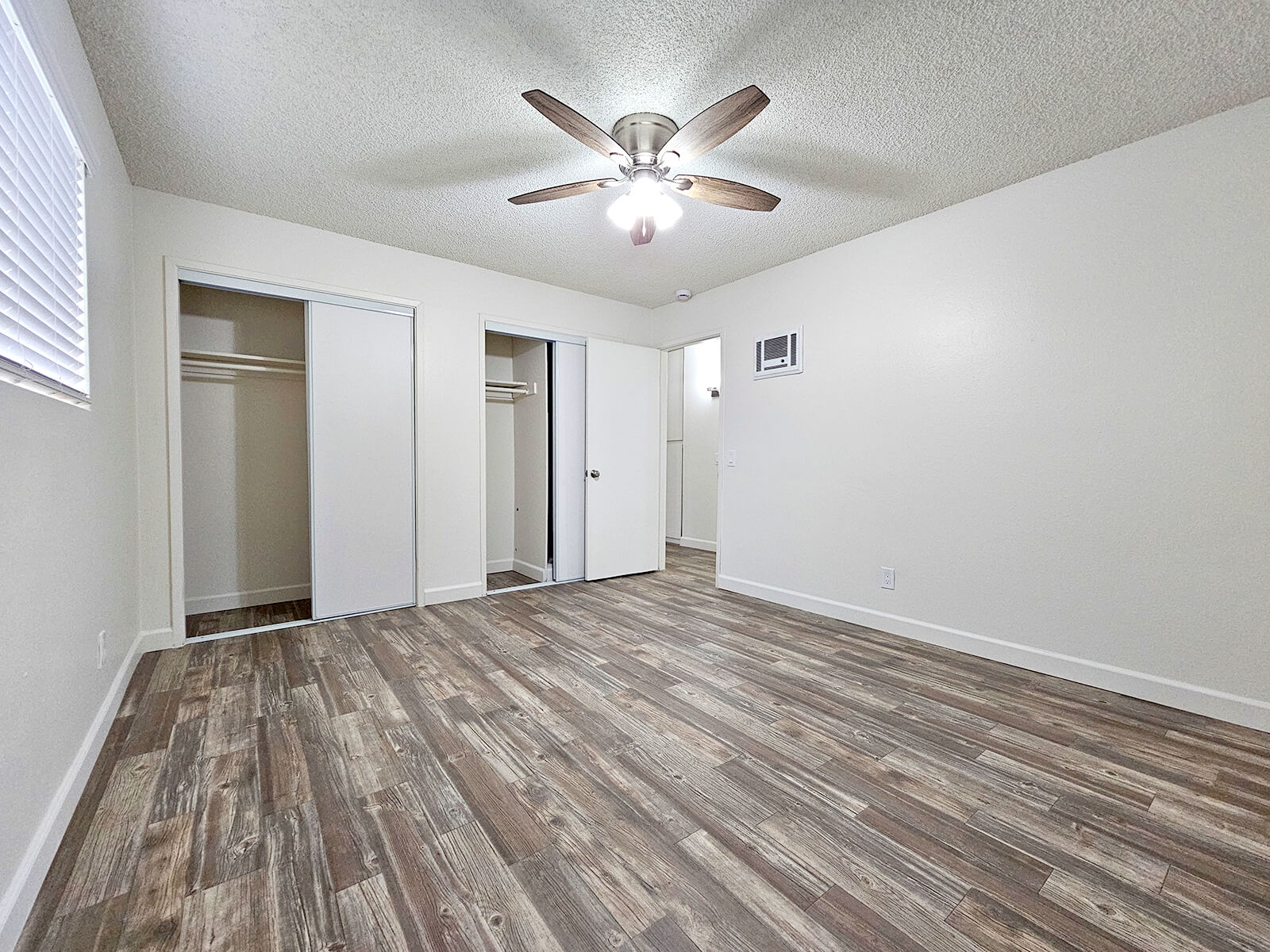 A spacious, unfurnished room featuring a ceiling fan, light wood-like flooring, and neutral-colored walls. Two closet doors are visible on the left, and a small vent is positioned near the ceiling. Natural light filters in through a window, enhancing the bright and airy atmosphere.