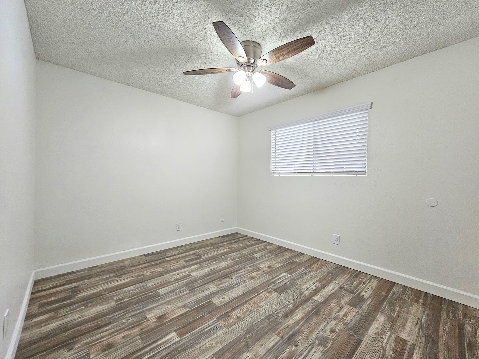 A clean, empty room with light-colored walls and a wooden floor. A ceiling fan with three lights is installed, and a window with white blinds allows natural light to enter. The room has no furniture, creating a spacious, minimalistic feel. The overall ambiance is bright and inviting.