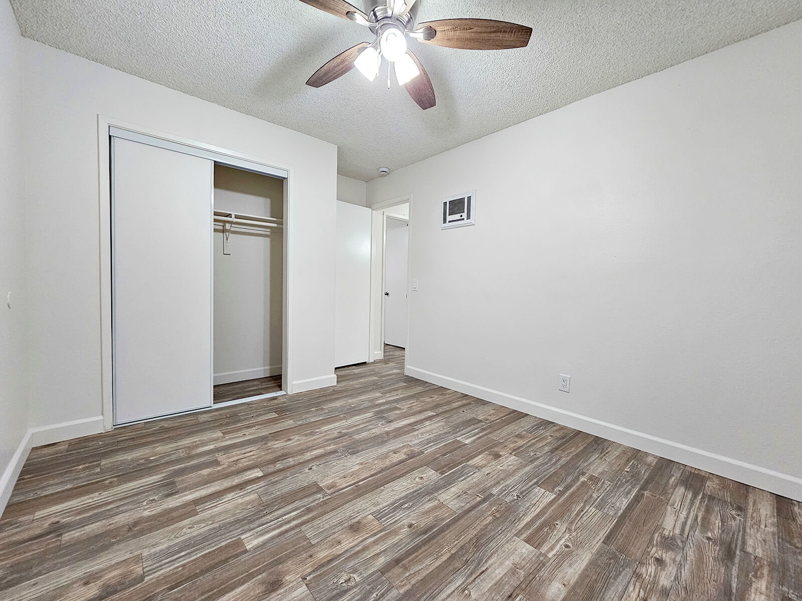 A spacious, empty room featuring light-colored walls, a ceiling fan with wooden blades, and laminate flooring. A sliding closet door is visible, along with a small wall-mounted air vent. The room is well-lit and has no furniture, providing a blank canvas for decoration.