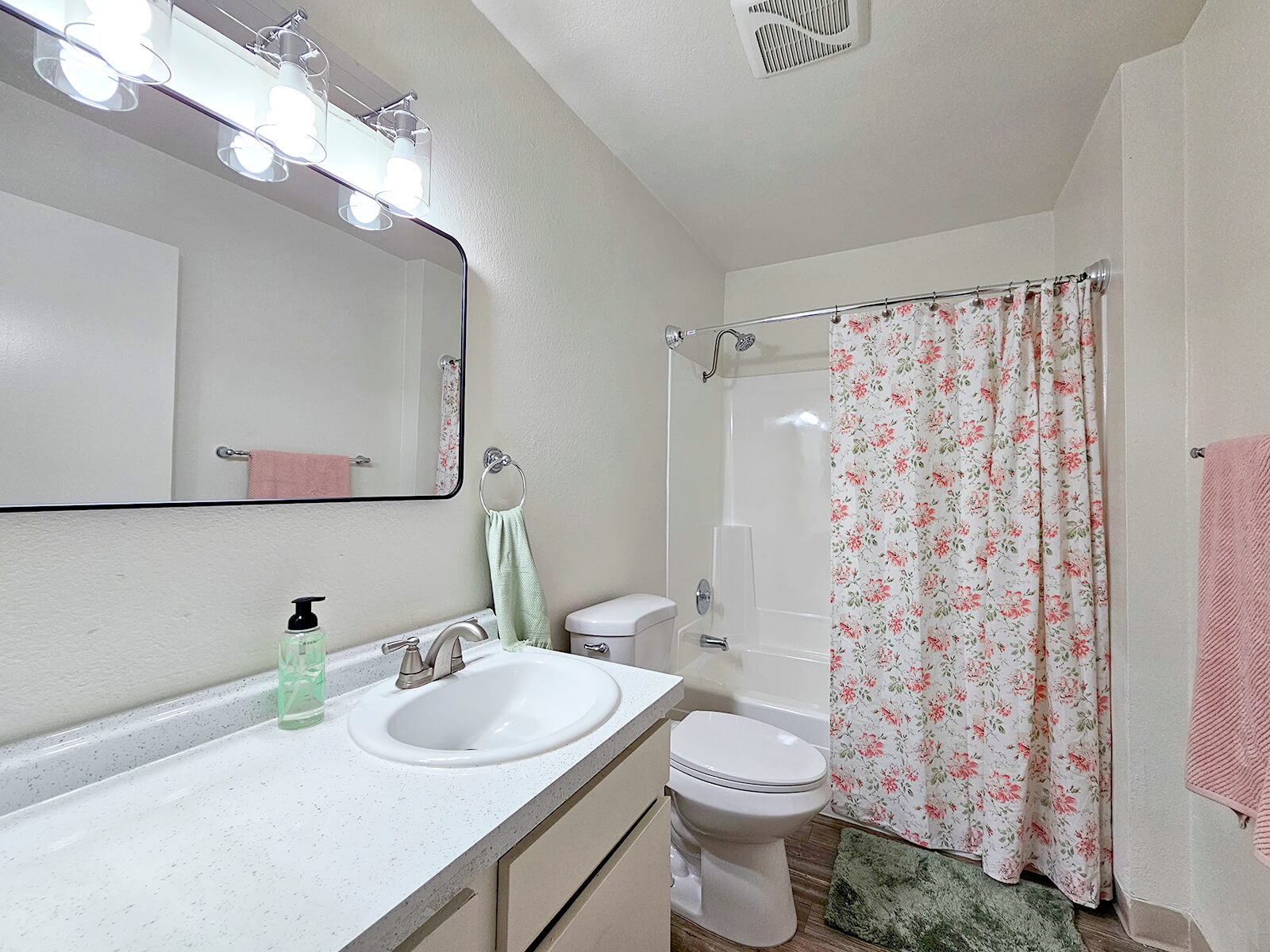 A clean bathroom featuring a white countertop with a sink, a large mirror above, a floral shower curtain, and a toilet. Soft pink towels hang on the wall, and a green bottle is placed on the counter. The walls are painted light colors, creating a bright and inviting atmosphere.
